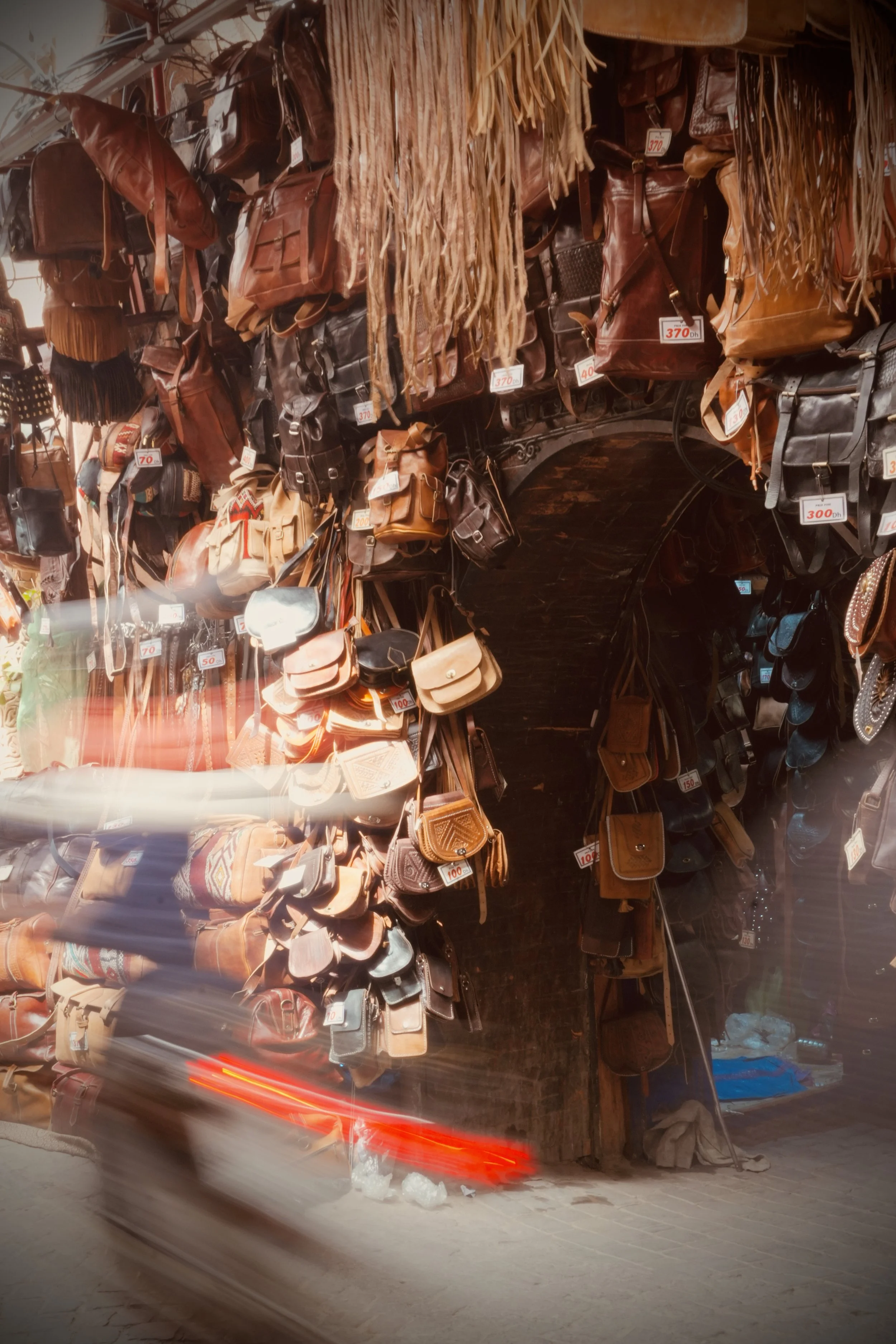 Un étal de marché avec de nombreux sacs en cuir suspendus, tagués avec des prix. On y voit des sacs à bandoulière, des sacs à dos et des sacs à main. Il y a également un scooter flou qui passe devant.