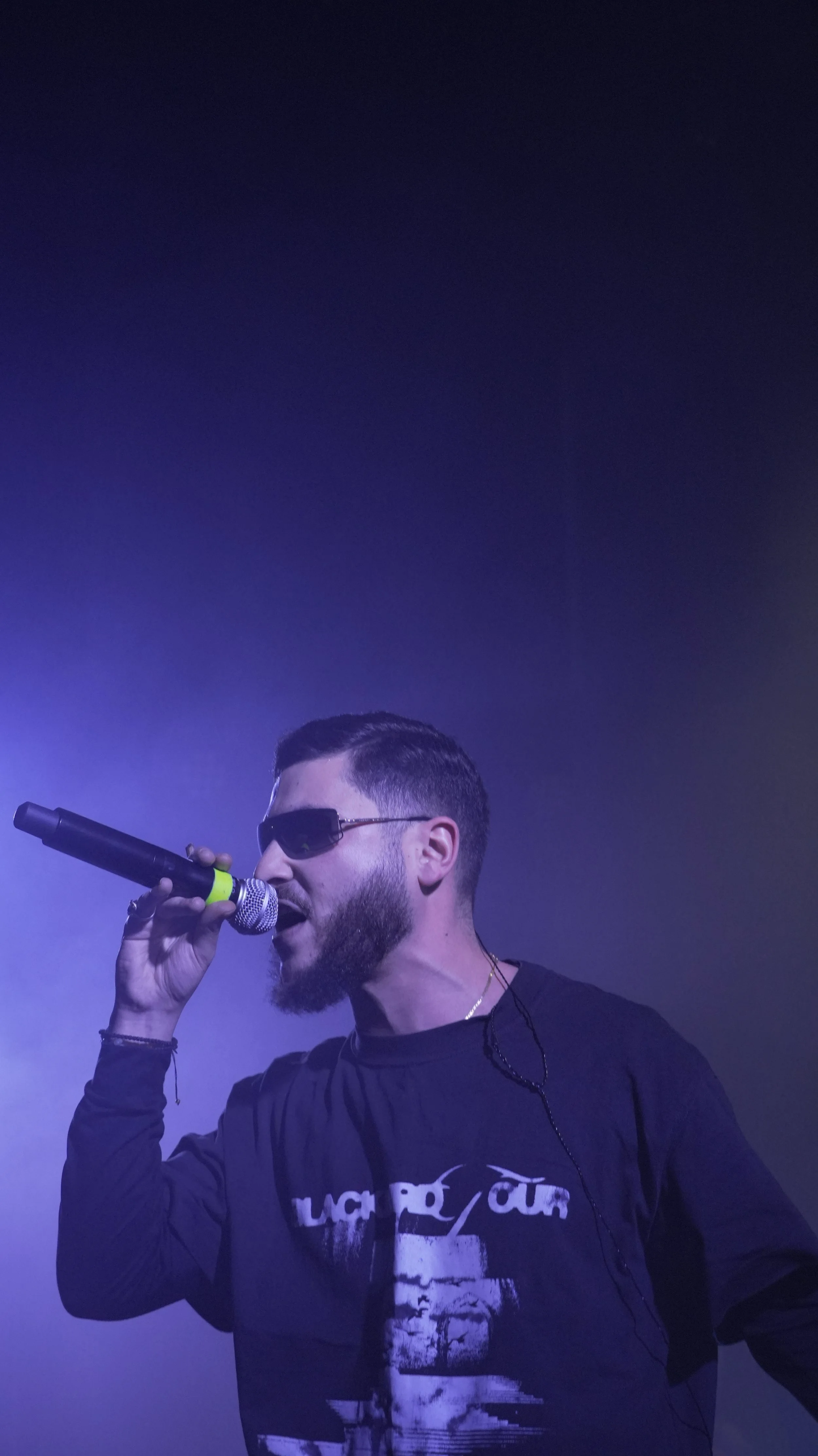 Chanteur avec lunettes de soleil et barbe, tenant un micro, sur scène avec éclairage violet.