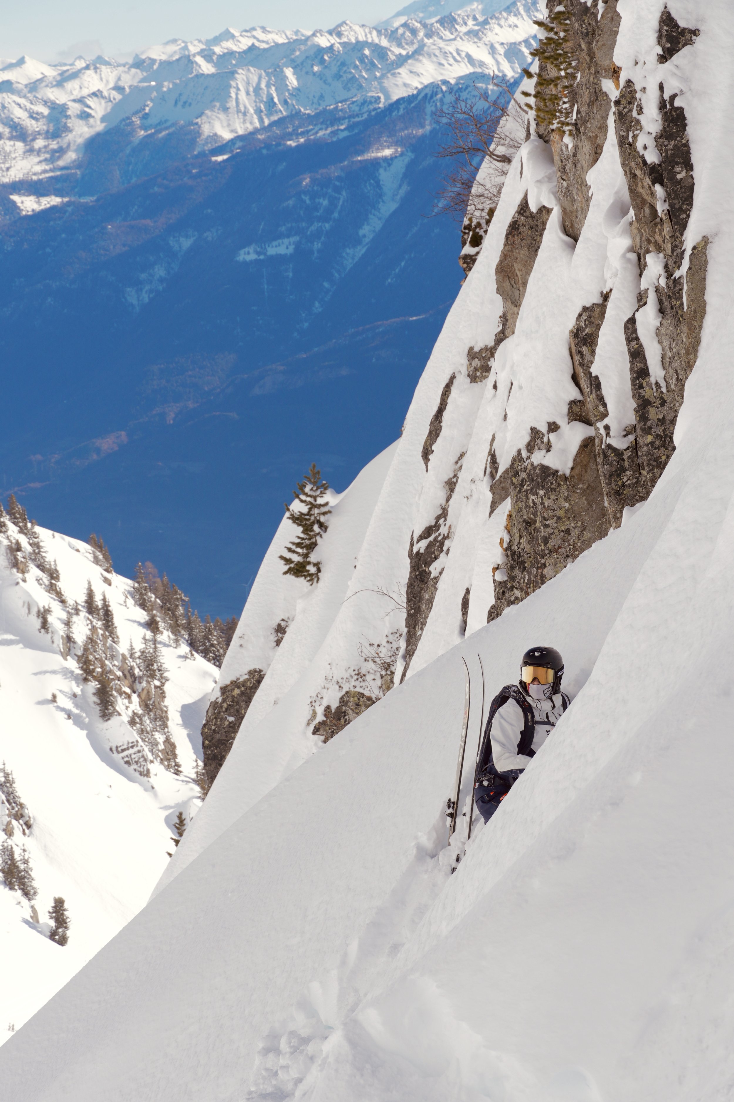 Skieur alpin escaladant une pente enneigée raide avec vue sur les montagnes, portant un casque et un masque de ski.