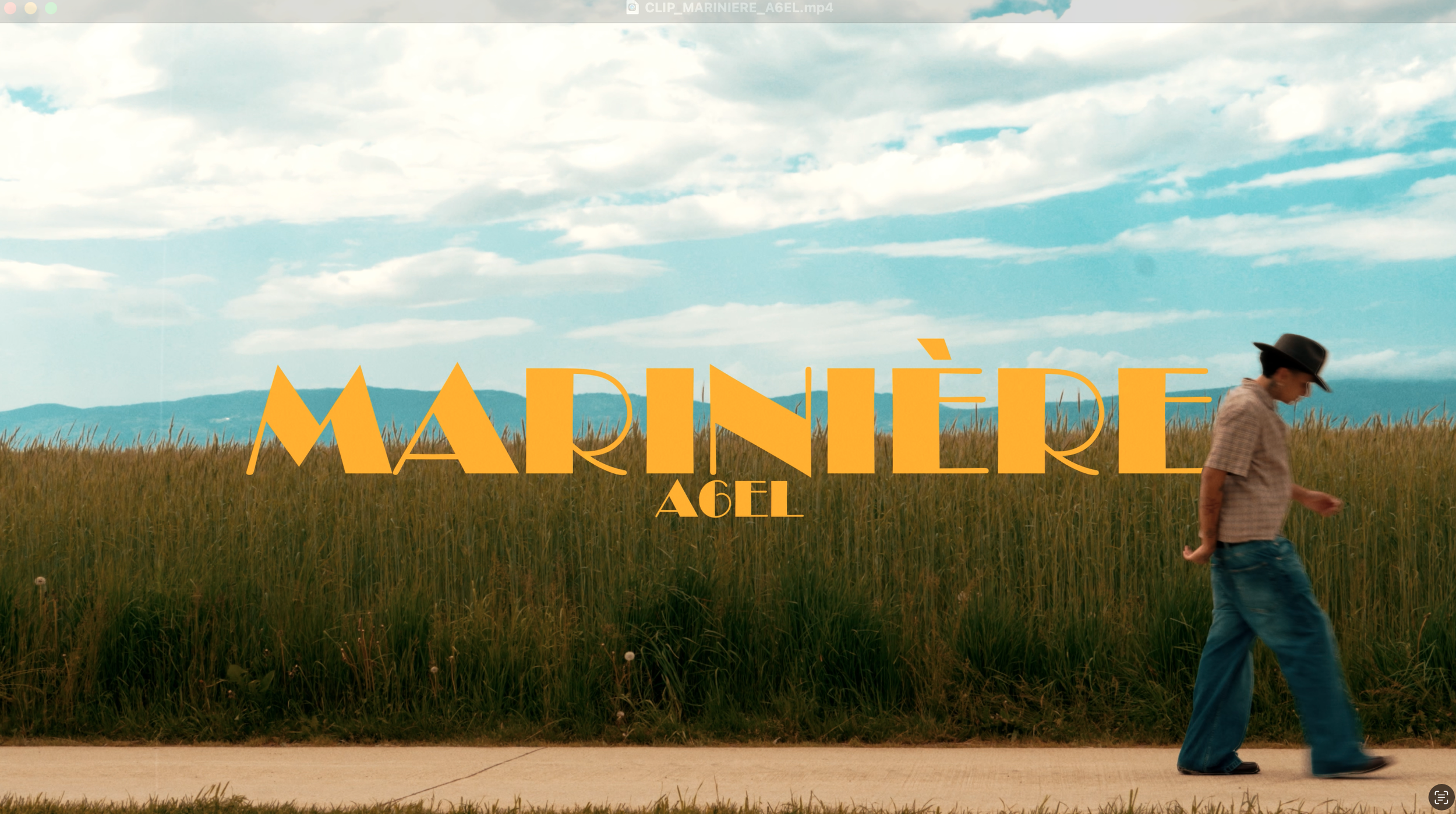 Une personne marchant près d'un champ sous un ciel bleu, avec le texte "MARINIÈRE A6EL" en lettres jaunes.