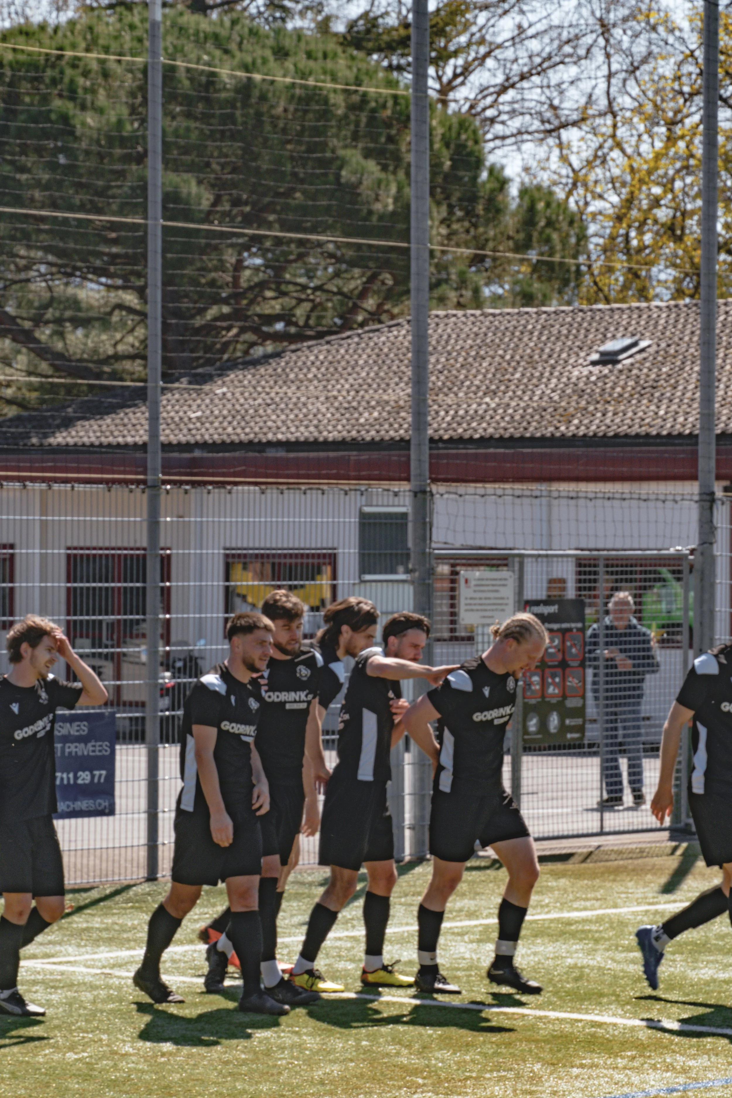 Un groupe de joueurs de football en uniformes noirs se tient sur un terrain, près d'une clôture métallique, pendant une journée ensoleillée.