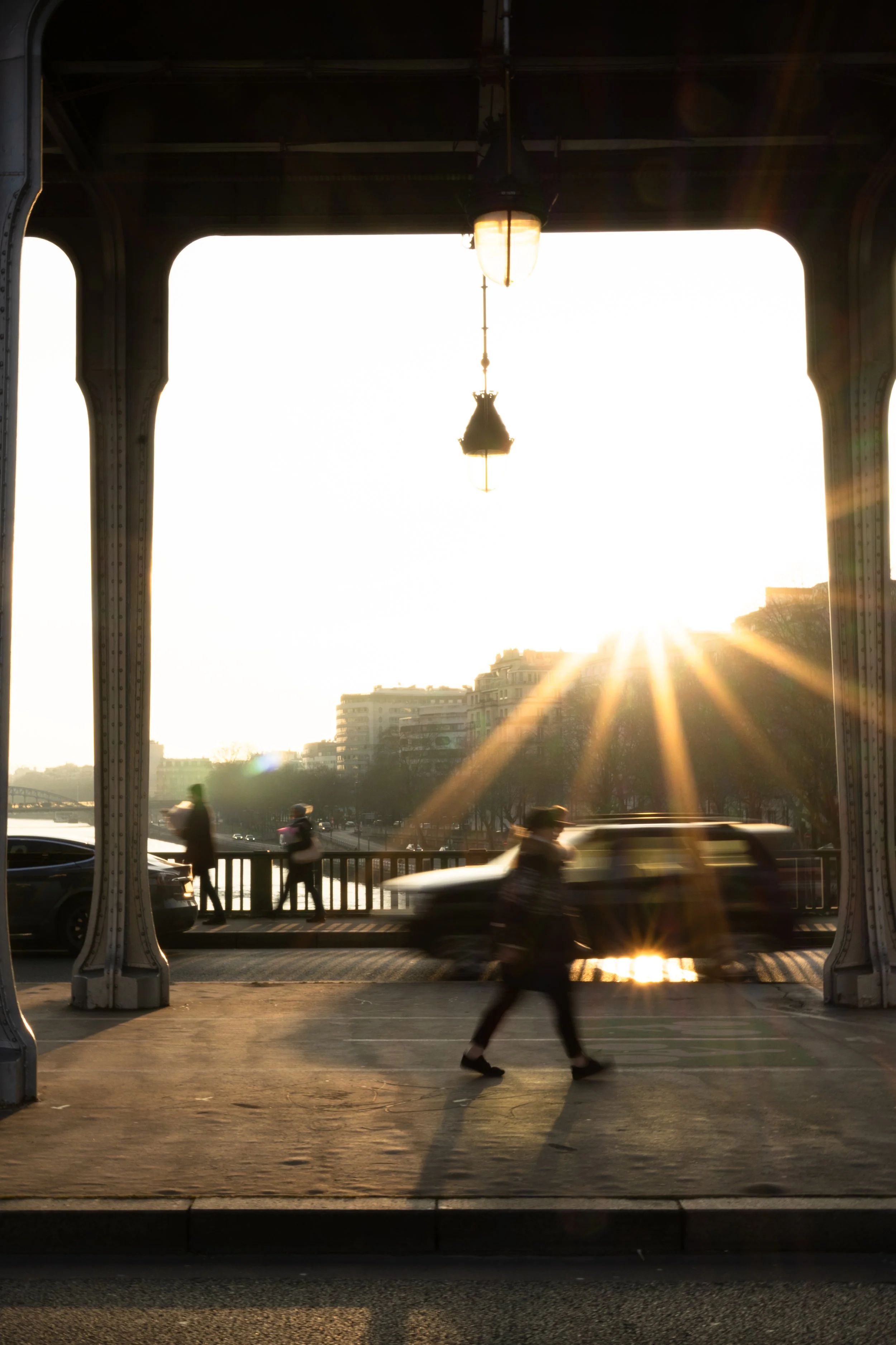 Vue d'un pont métallique au coucher du soleil, avec des silhouettes de piétons et de voitures, éclairées par les rayons du soleil.