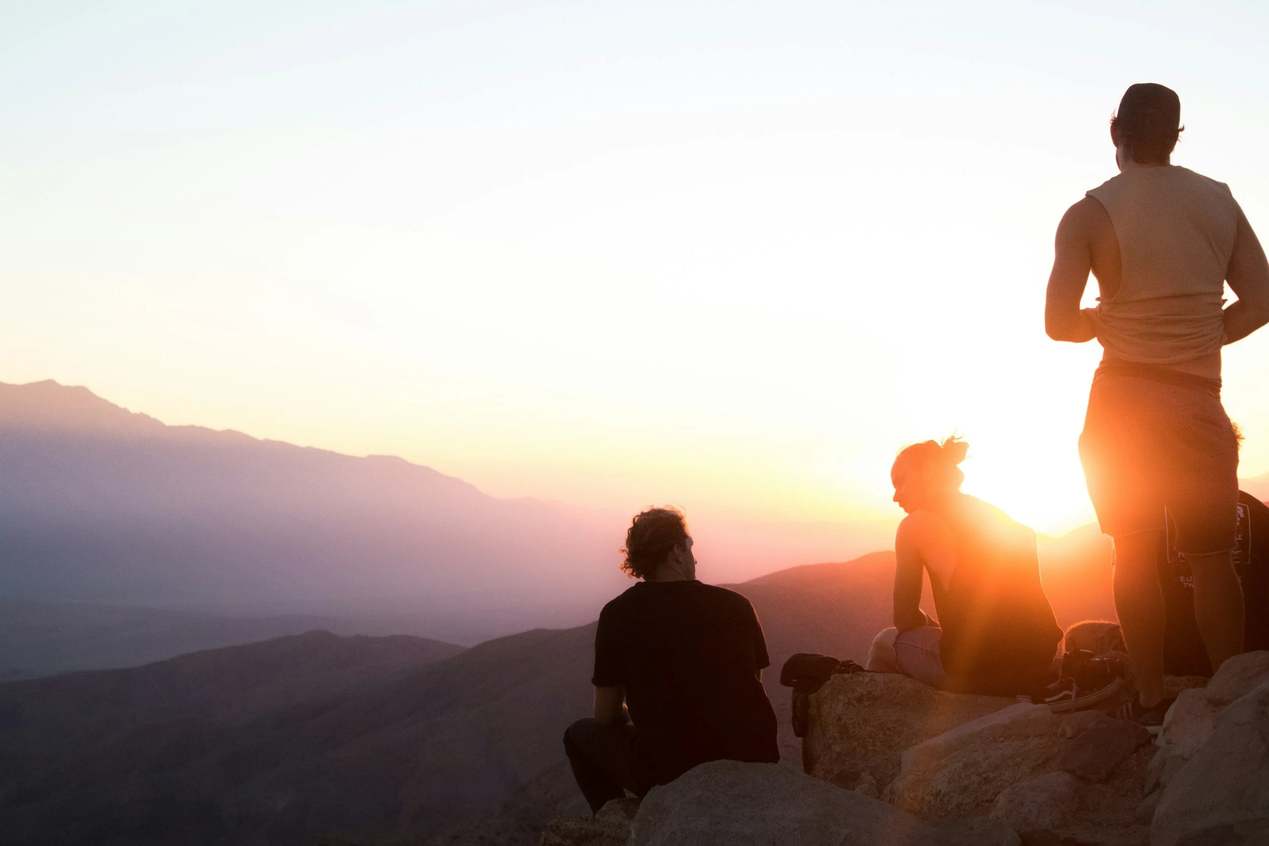 Four people sitting and standing on rocks during sunset in a mountainous landscape.