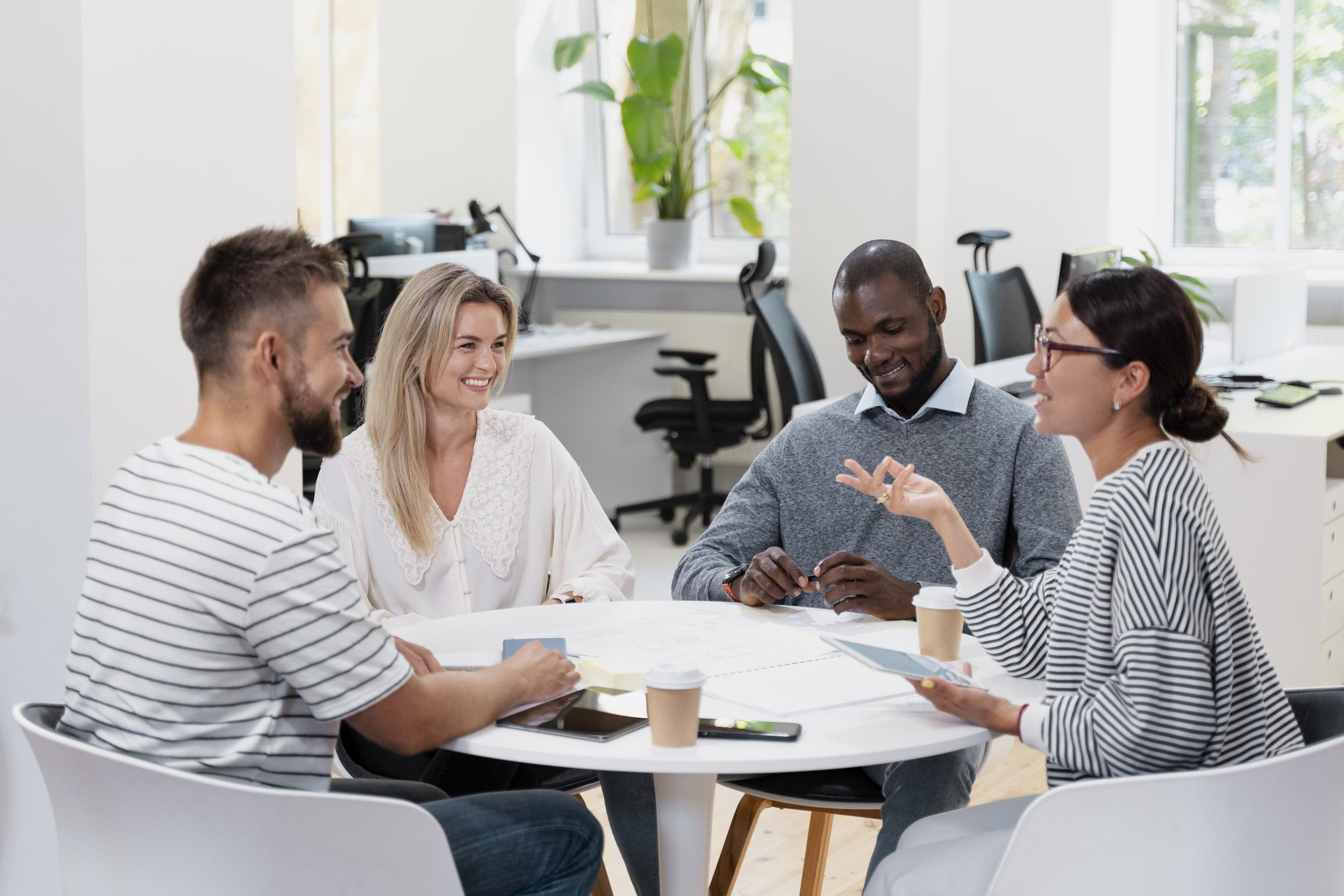 Four diverse colleagues smiling and talking during a meeting in a bright office with large windows and green plants.