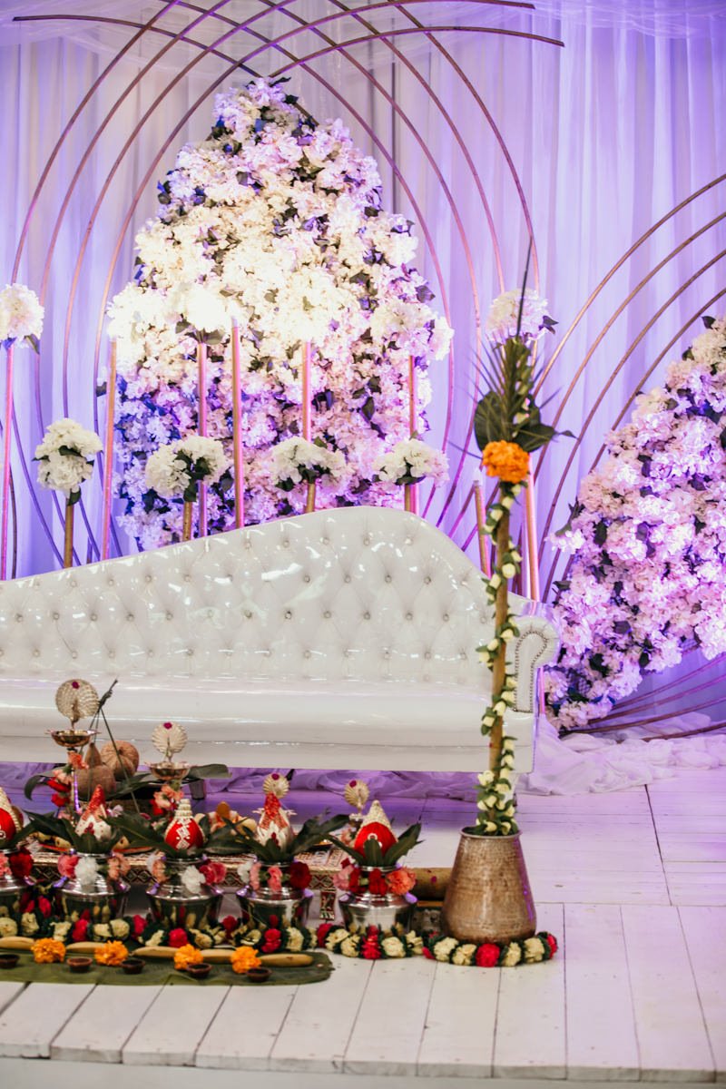 A decorated stage with a white tufted sofa, surrounded by large floral arrangements of white and purple flowers, and traditional Indian wedding items including kalash and decorations.