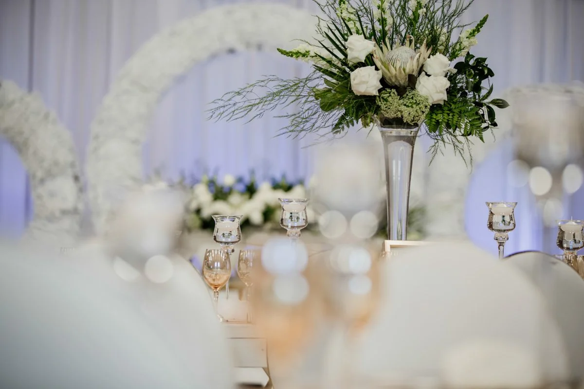 Elegant banquet table setup with a tall floral centerpiece, candles, and a backdrop of white drapes for a special event.