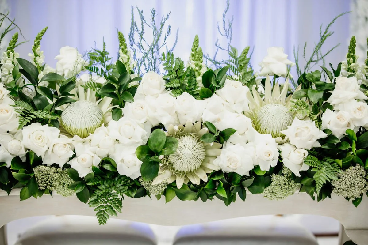 A floral arrangement with white roses, green leaves, and textured white flowers on a white table with a blurred white curtain background.