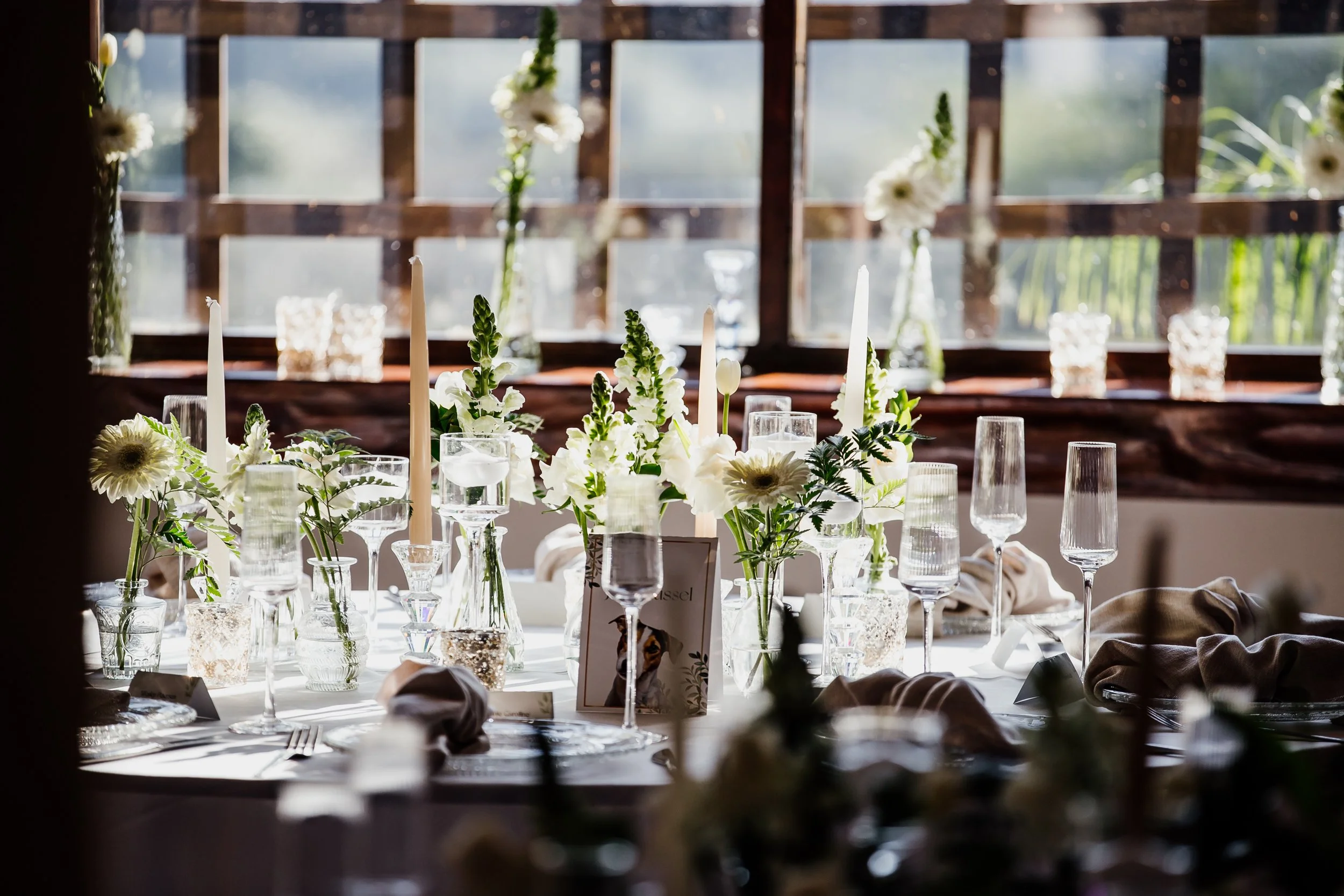Elegant table setting with white flowers, candles, and glassware at a wedding or special event, illuminated by natural light from a window.