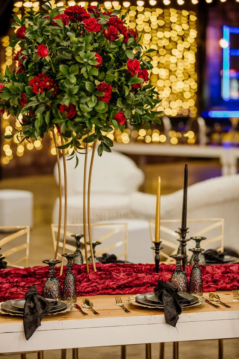 Elegant dining table with black plates, gold utensils, and black napkins, decorated with tall gold candlesticks and a large floral centerpiece of red roses and greenery, set against a background of warm string lights and cozy furniture.