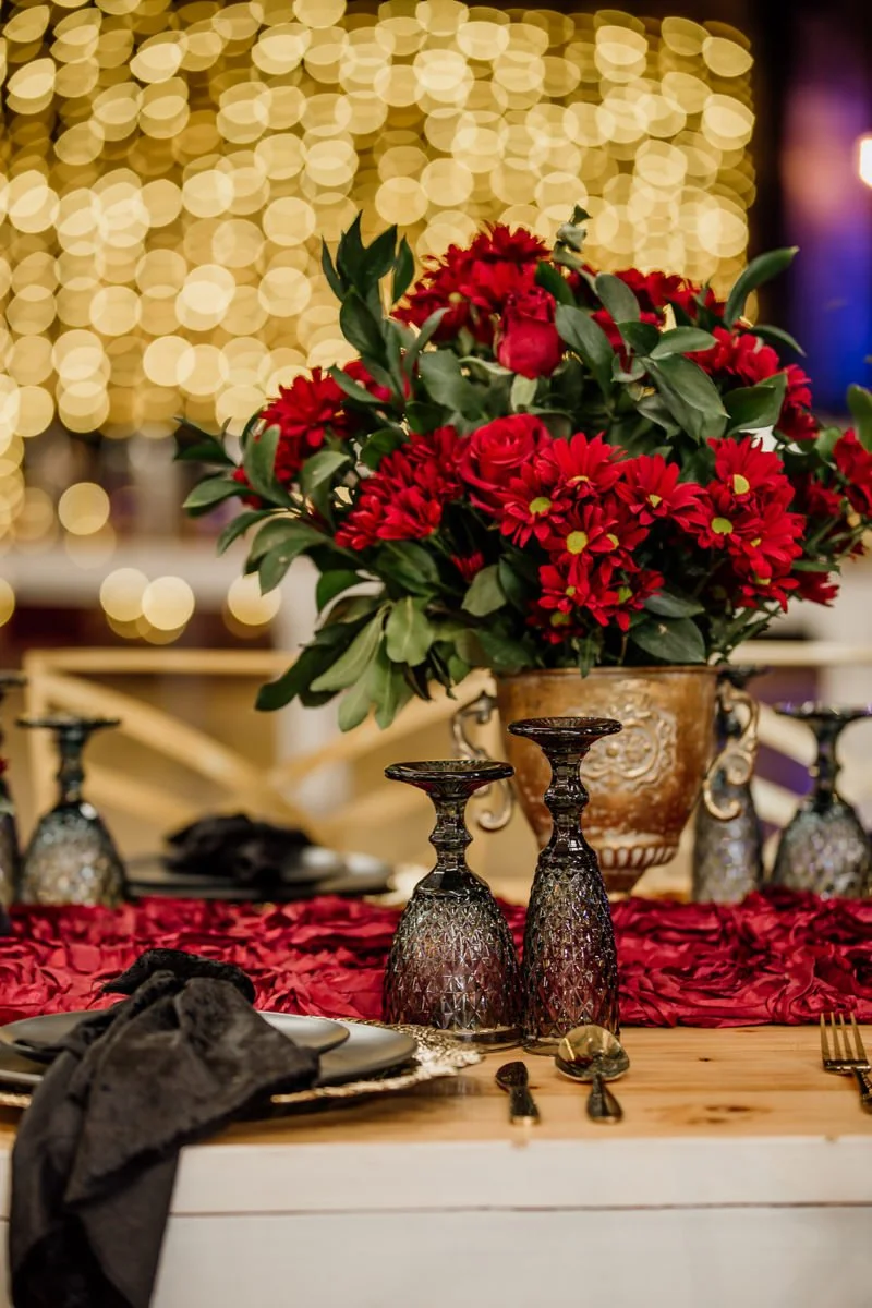 A festive dining table decorated with a large bouquet of red flowers in a gold vase, surrounded by ornate glass candle holders, black napkins, and golden cutlery, with warm string lights creating a cozy ambiance.