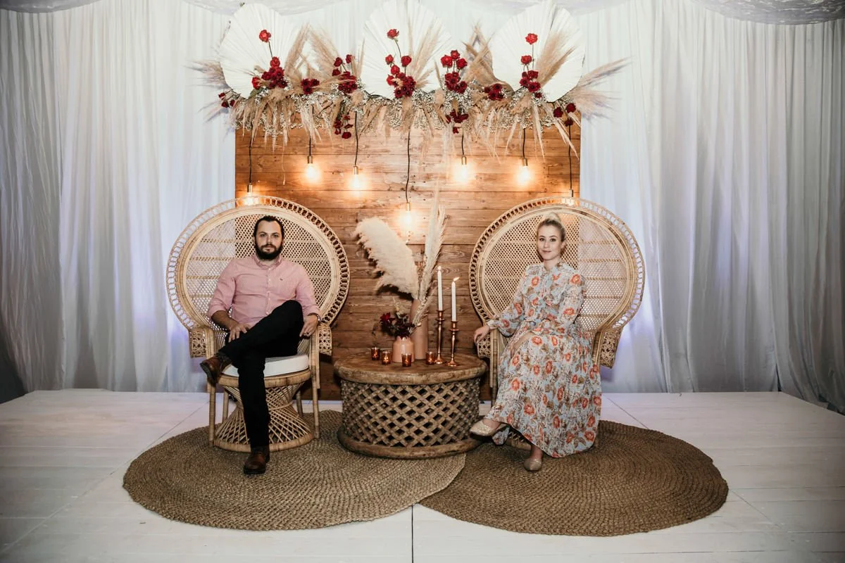 Two people sitting in wicker chairs on circular rugs, with a wooden backdrop decorated with floral arrangements and hanging lights.