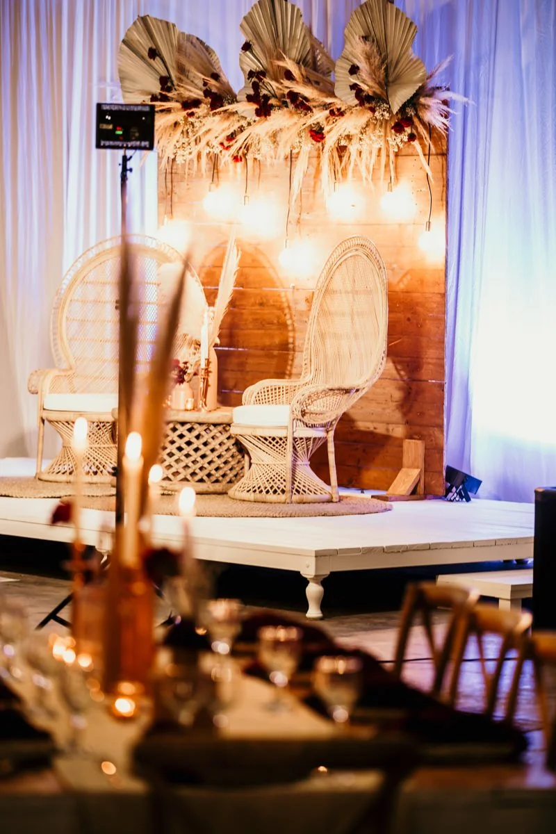Boho wedding or event stage with wicker chairs, wooden backdrop decorated with dried palm leaves and flowers, string lights, and curtains, with a table's edge and candles in the foreground.