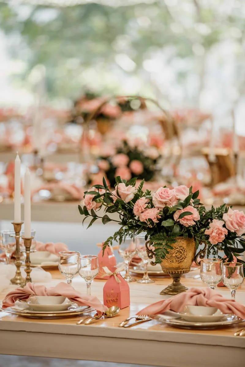 Elegant dining table set for a wedding or celebration, decorated with pink roses, glassware, gold utensils, pink napkins, and white candles in gold holders.