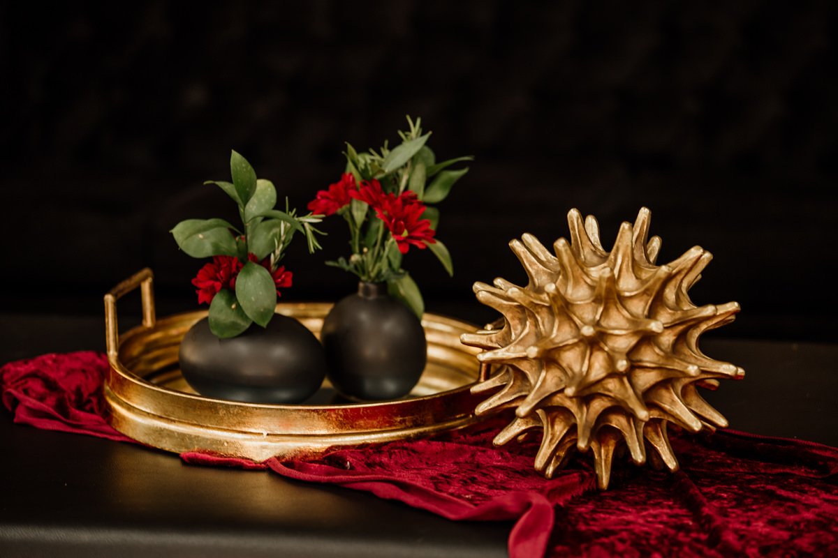 Decorative arrangement with two black vases of greenery and red flowers, a textured gold-colored object, all on a gold tray on a dark background with red cloth underneath.
