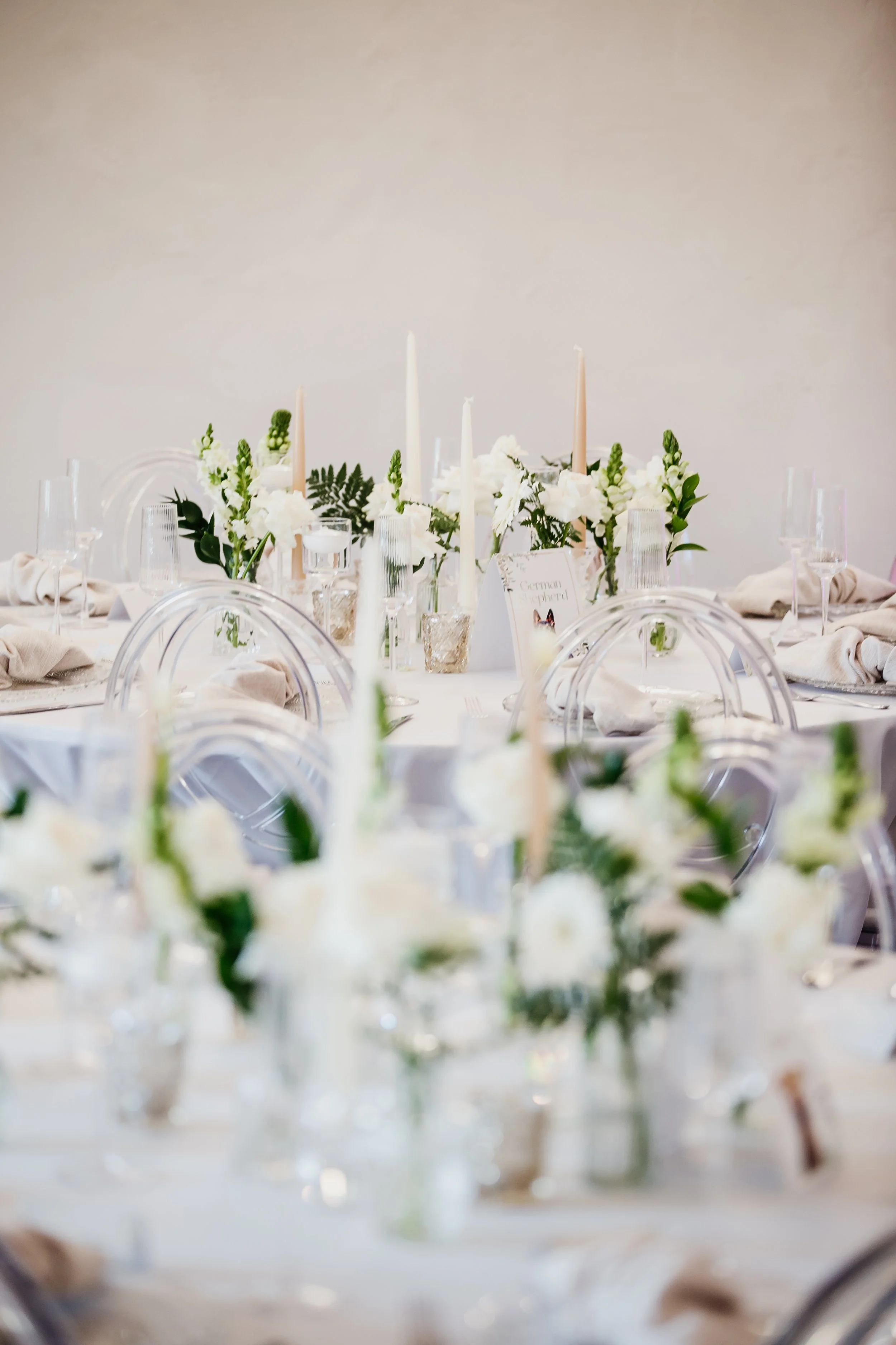 Elegant wedding reception table with white and green floral arrangements, white candles, and clear glassware and chairs.