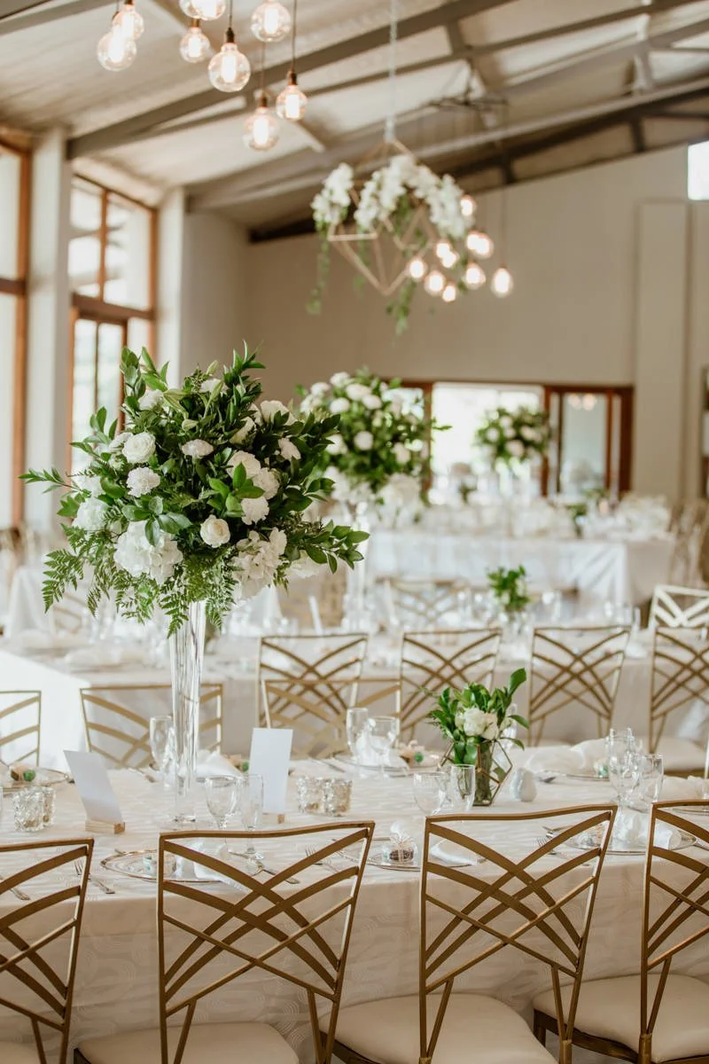 Elegant wedding reception decorated with white floral centerpieces and gold chairs in a bright, airy venue with wall-to-wall windows and hanging lights.