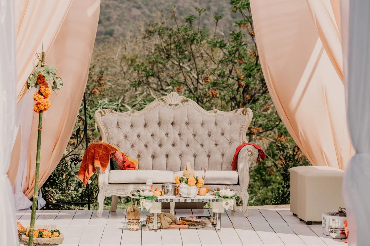 A vintage, ornate sofa with tufted upholstery on a decorated outdoor patio, surrounded by pink curtains, with a small table holding flowers and pumpkin decorations, set against a backdrop of trees and foliage.