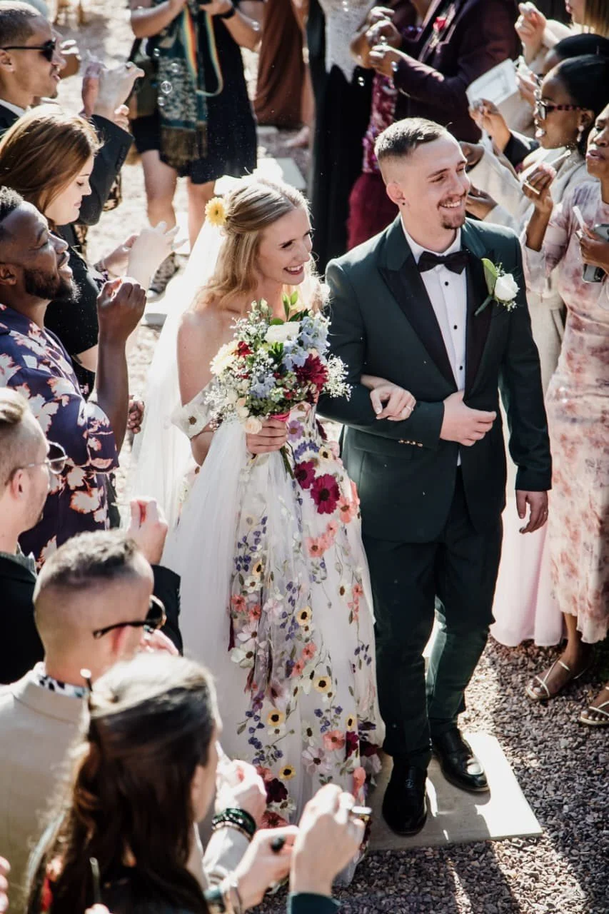 A bride and groom smiling and walking together surrounded by guests at an outdoor wedding.