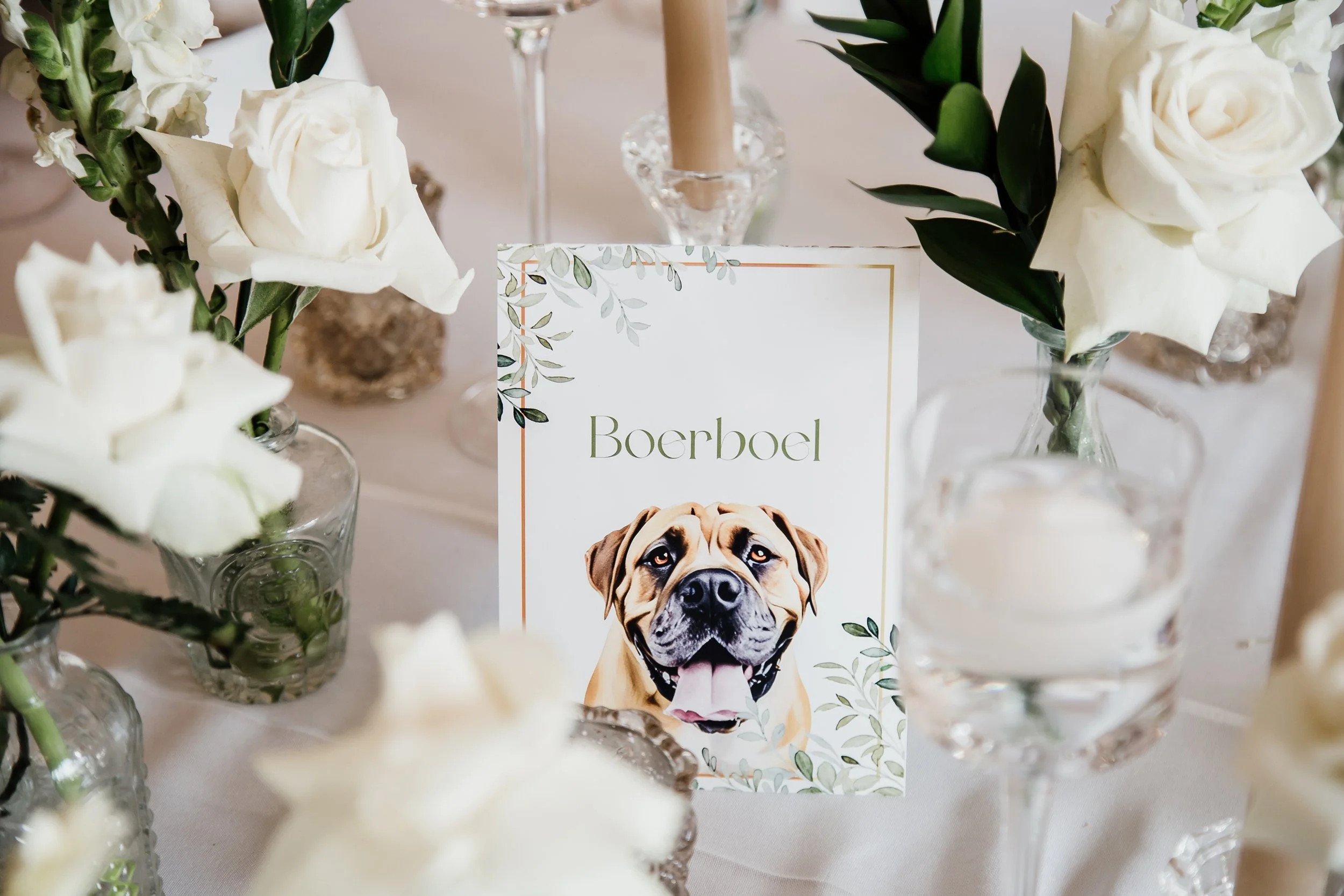 Table centerpiece with white roses and greenery, decorative candles, and a printed card featuring a happy dog with the name 'Boerboel'.