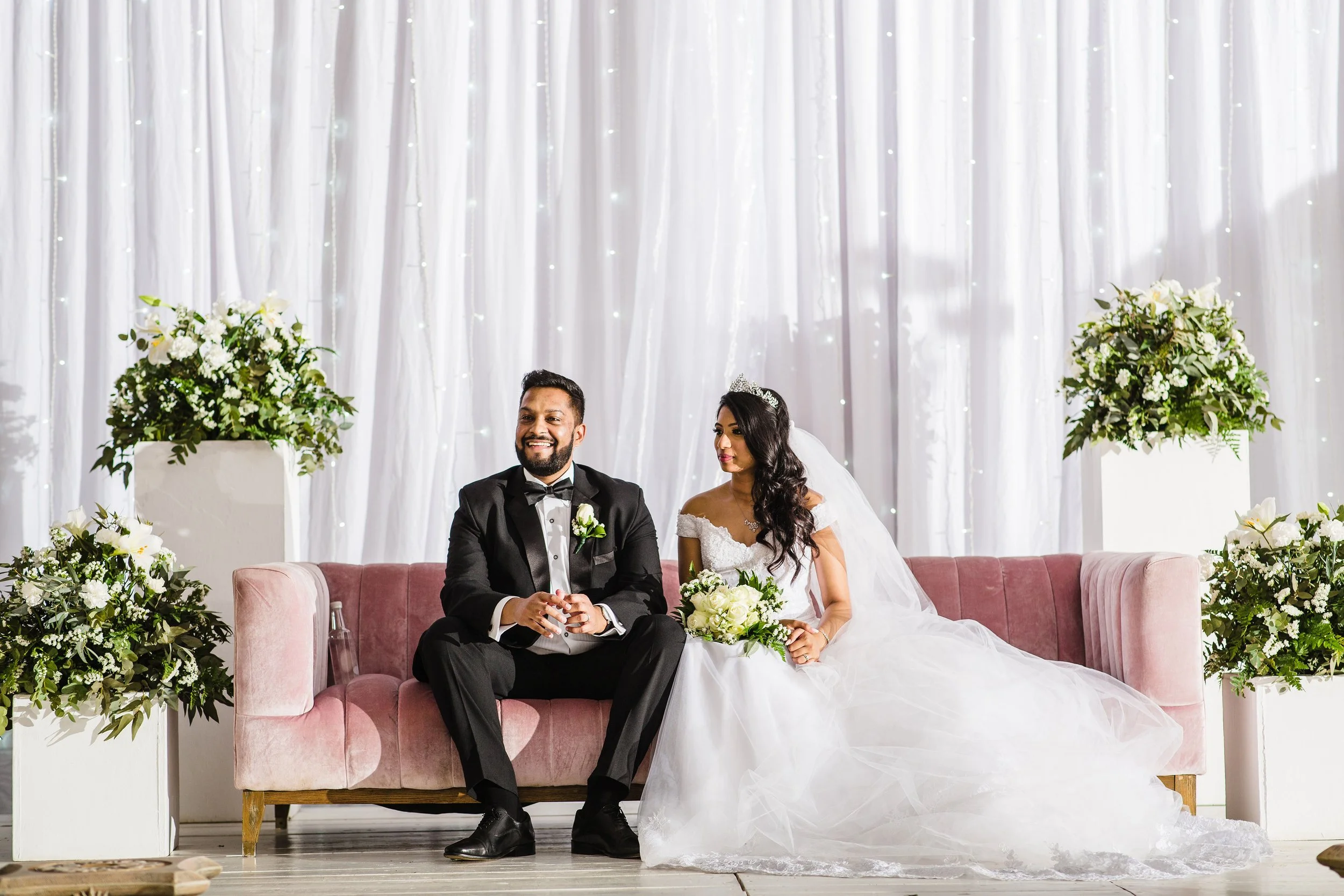 Bride and groom sitting on a pink couch during a wedding ceremony, with white drapes and floral arrangements in the background.