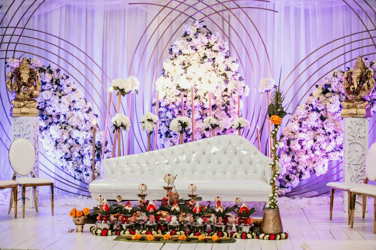 Decorated stage with white tufted seating, floral arrangements, and Lakshmi statues, likely for a Hindu wedding or religious ceremony.
