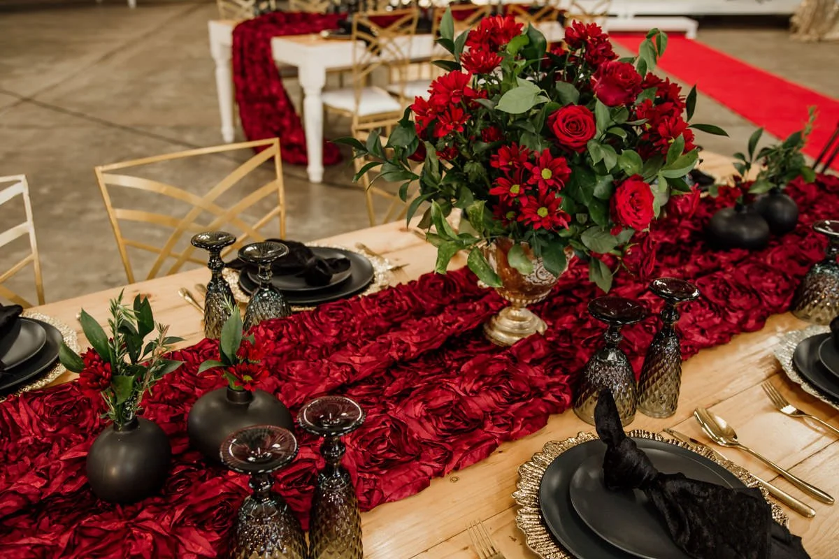 A decorated dining table featuring a red floral centerpiece, black plates with silver chargers, black napkins, gold and black glassware, and a red rose petal table runner. Gold chairs surround the table.