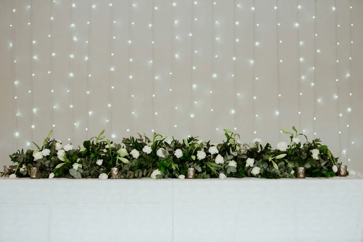 A decorated table with a floral arrangement of white flowers and green foliage, set against a backdrop of hanging string lights.