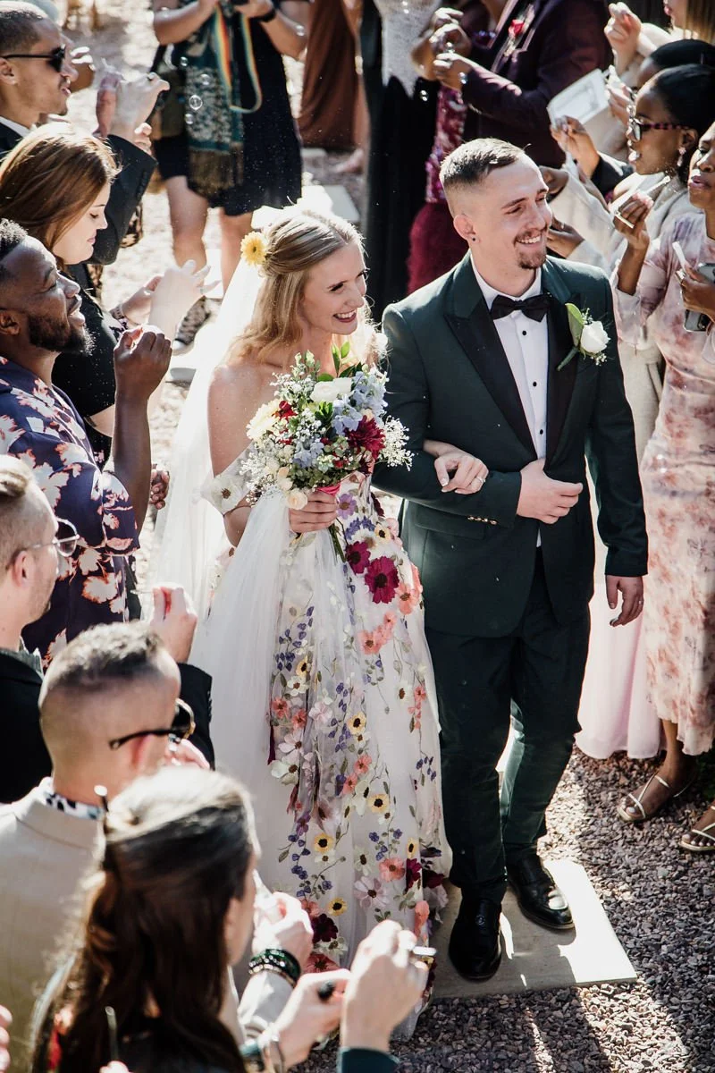 A bride and groom walking down the aisle surrounded by friends and family at an outdoor wedding.