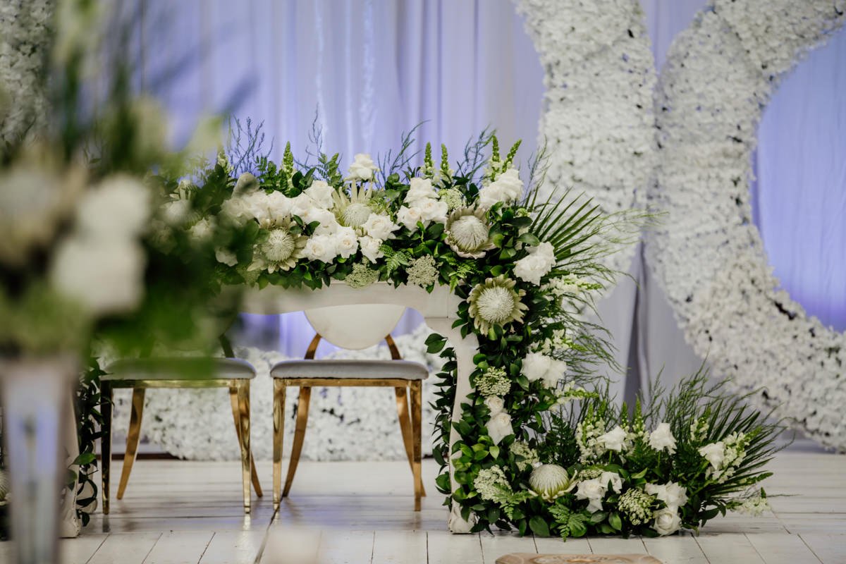 Decorative floral arrangement with white flowers and green foliage on and around a white table in front of a white floral arch.