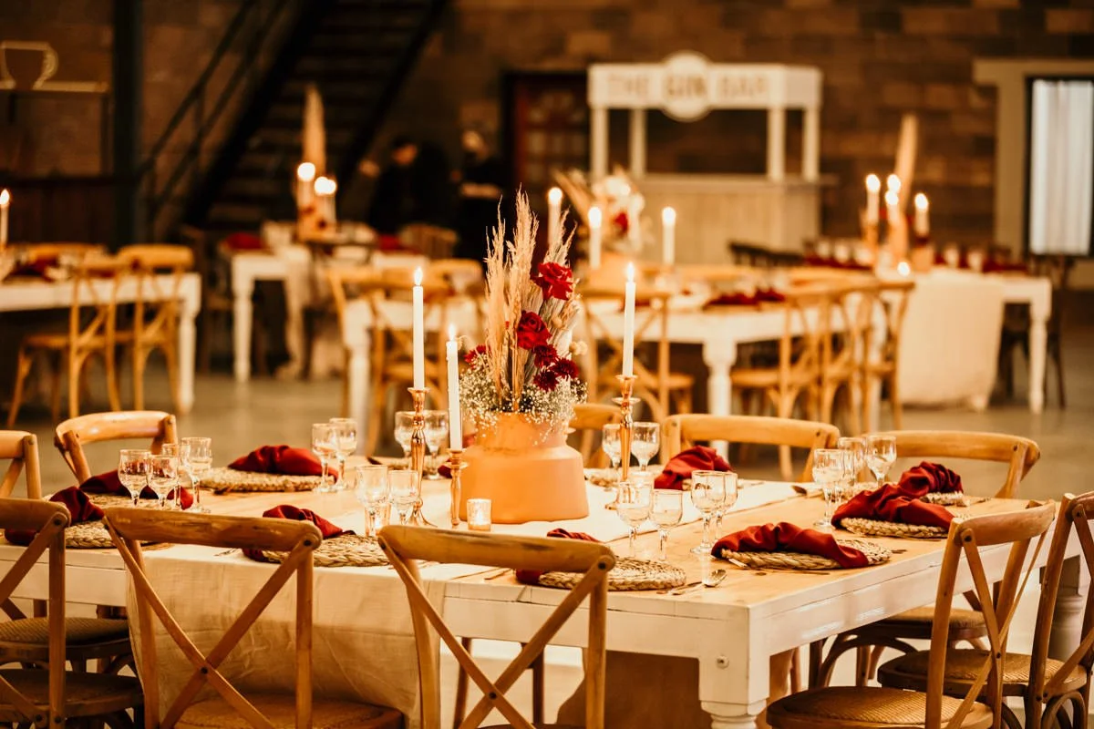 A decorated banquet table set for an event in a rustic style with beige tablecloths, red napkins, and glassware. A large floral centerpiece with red flowers and tall dried grasses is on the table, illuminated by candlelight. Wooden chairs surround th