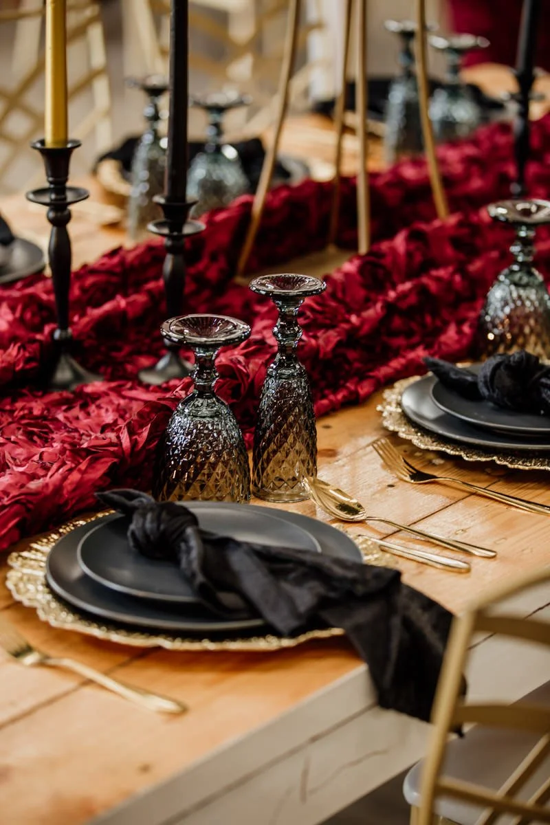 Elegant dining table set with black plates, black cloth napkins, gold flatware, black and gold textured glasses, black candlesticks, and a red floral table runner with gold accents.