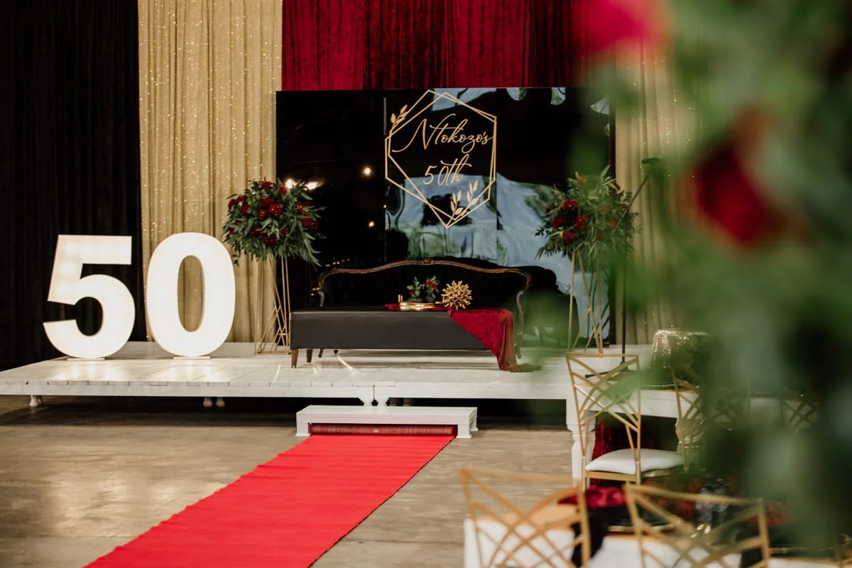 A decorated stage for a celebration, featuring large illuminated numbers '50', a black sofa with a red cloth, floral arrangements, and a backdrop with gold and black curtains and a sign that reads 'Nokozo's 50th'.