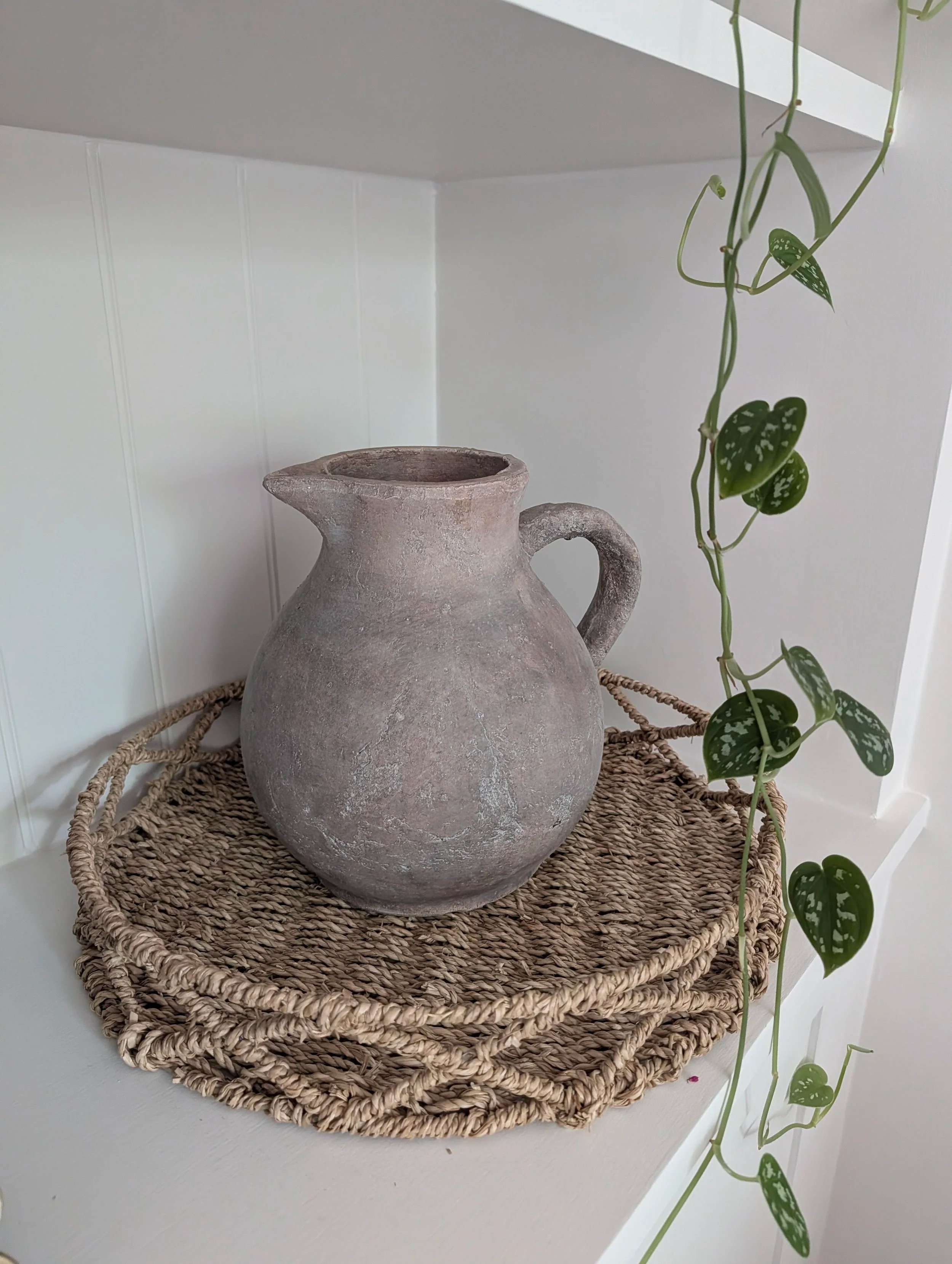 A rustic, weathered ceramic vase with a single handle is placed on a woven basket tray, with trailing green and white variegated heart-shaped leaves cascading nearby, all set against a white wall.
