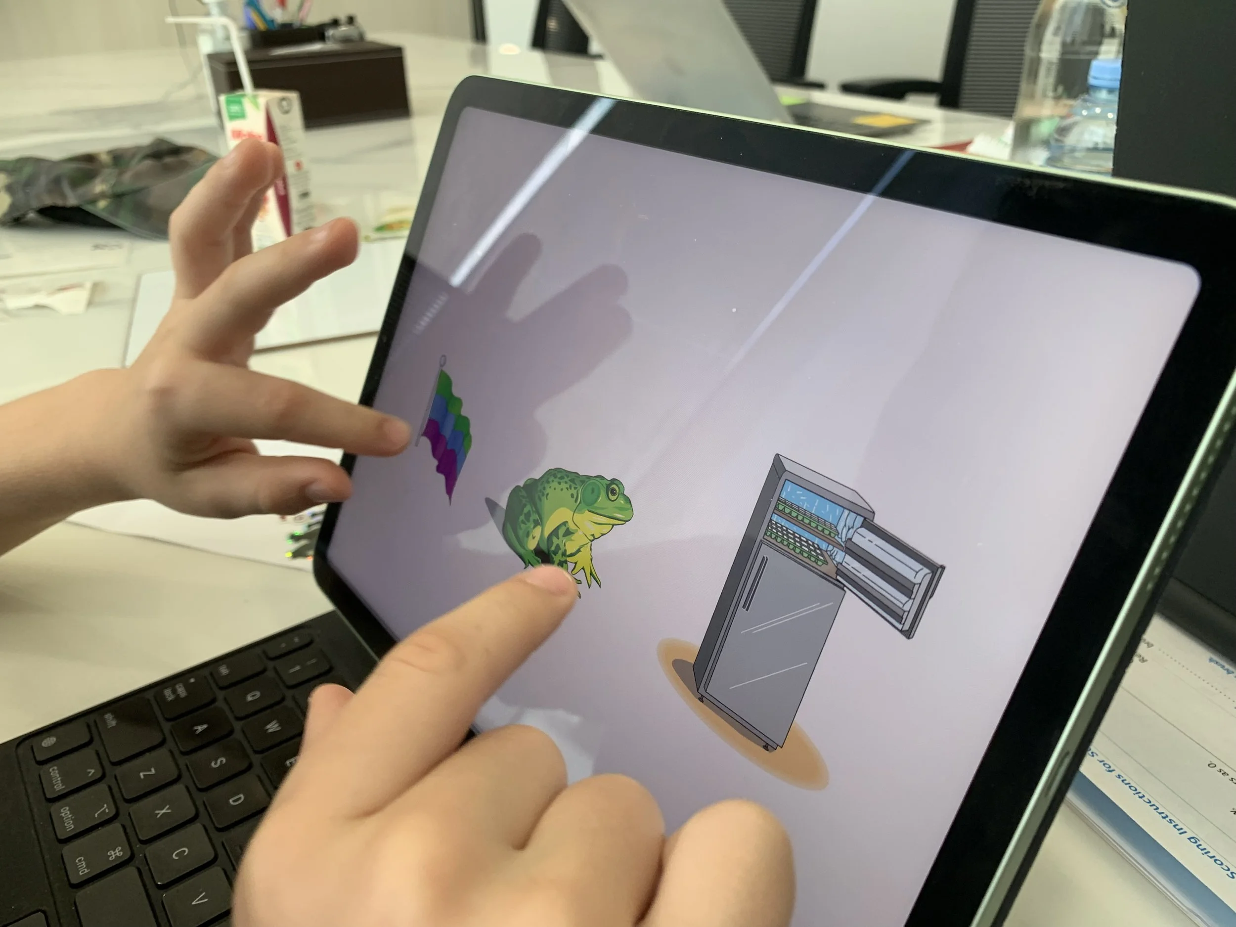 Child interacting with a tablet screen displaying images of colorful blocks, a frog, and a refrigerator, with a keyboard and desk items visible in the background.