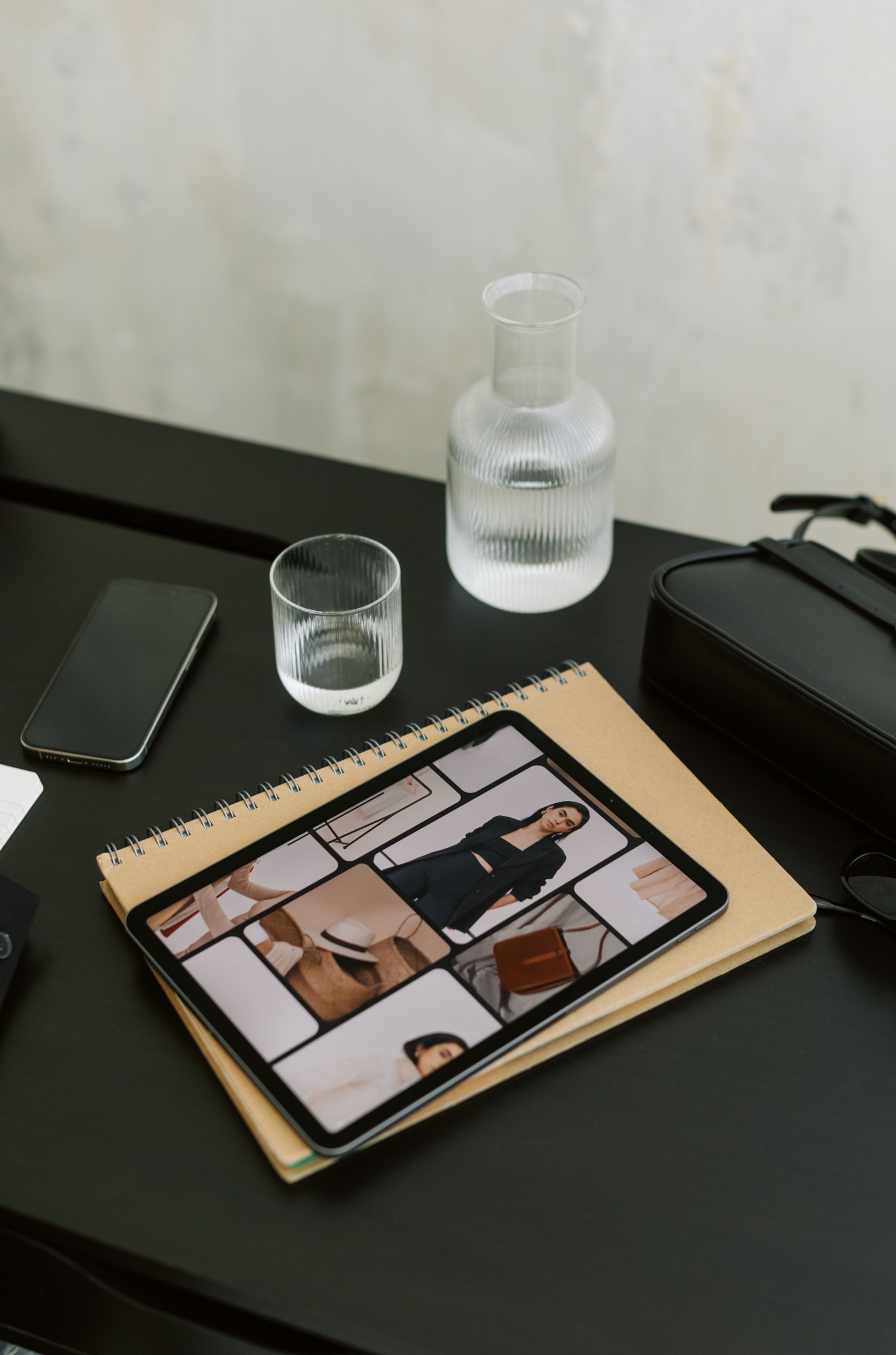 Tablet displaying fashion images on a spice notebook, next to a phone, glass of water, carafe, and black case on a dark desk.