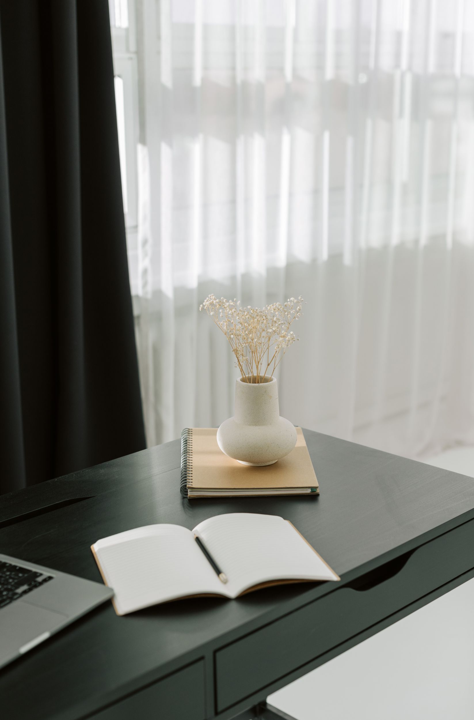 Minimalist workspace with dark desk, open notebook, laptop, and small vase with dried flowers; light curtains in background.