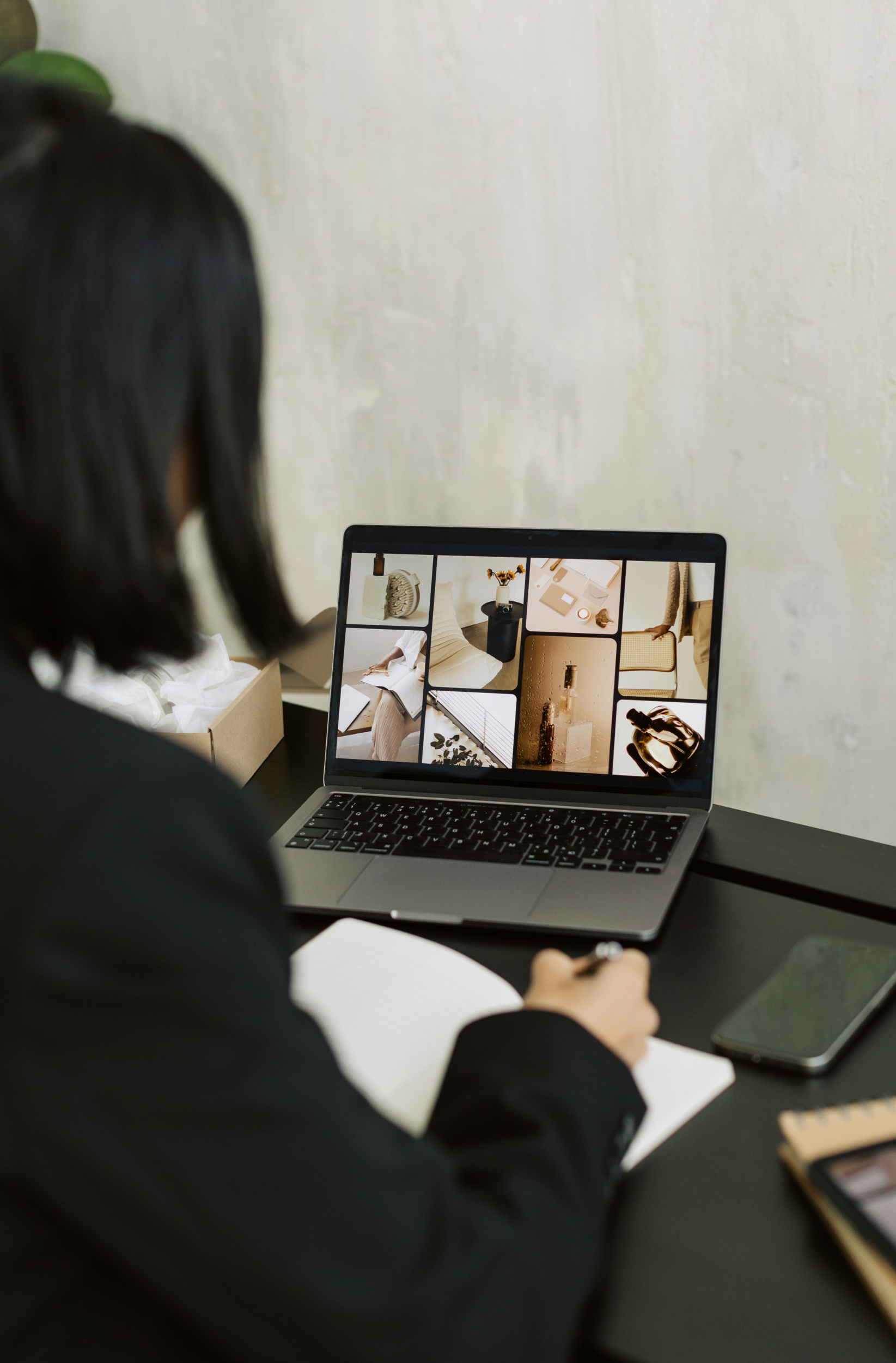 Person viewing mood board on laptop screen, seated at desk with notepad and smartphone.