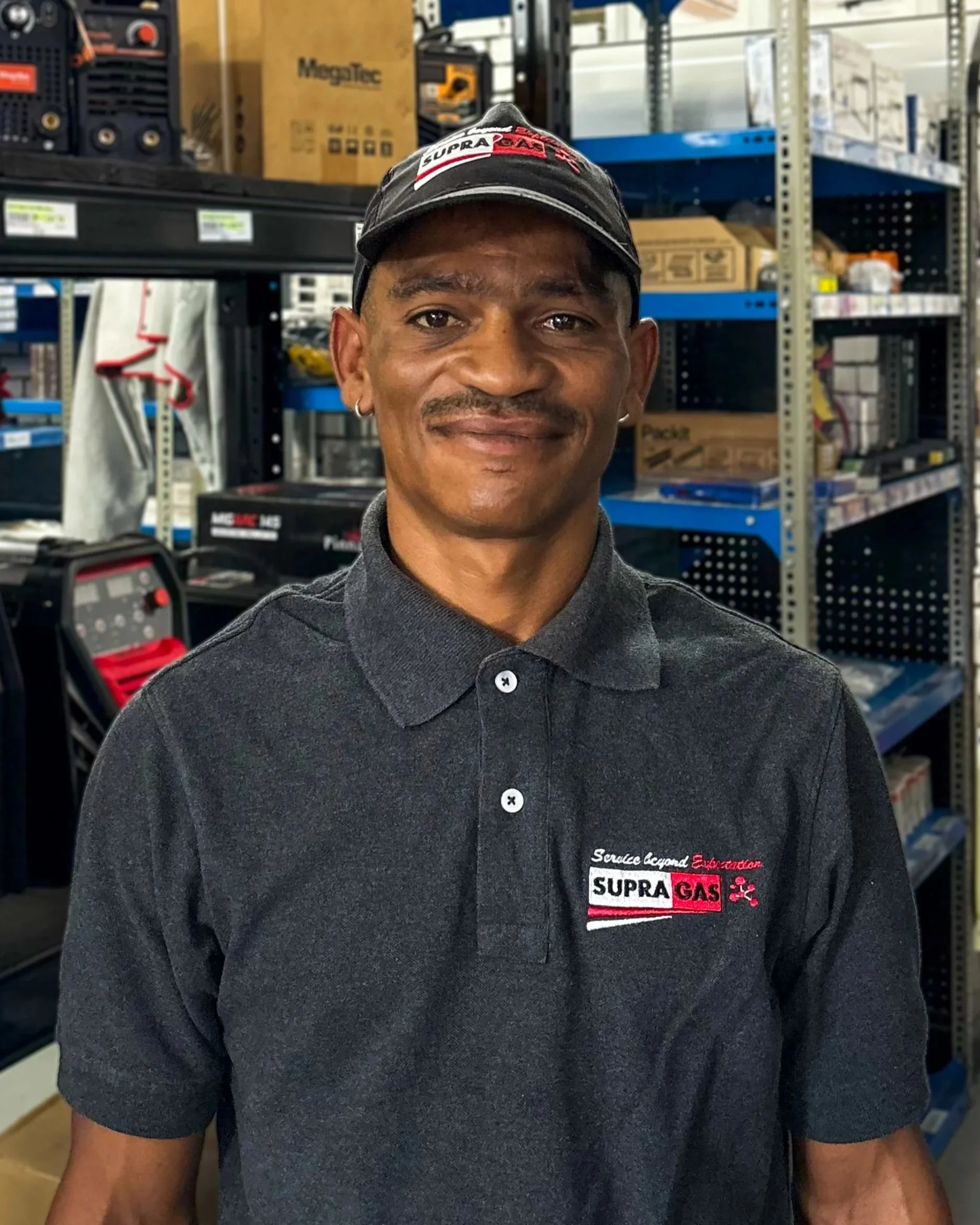 Delivery driver's assistant at Supragas. A man wearing a dark gray shirt and a black cap with red and white logos, standing in front of shelves in a hardware store aisle with various boxes and equipment.