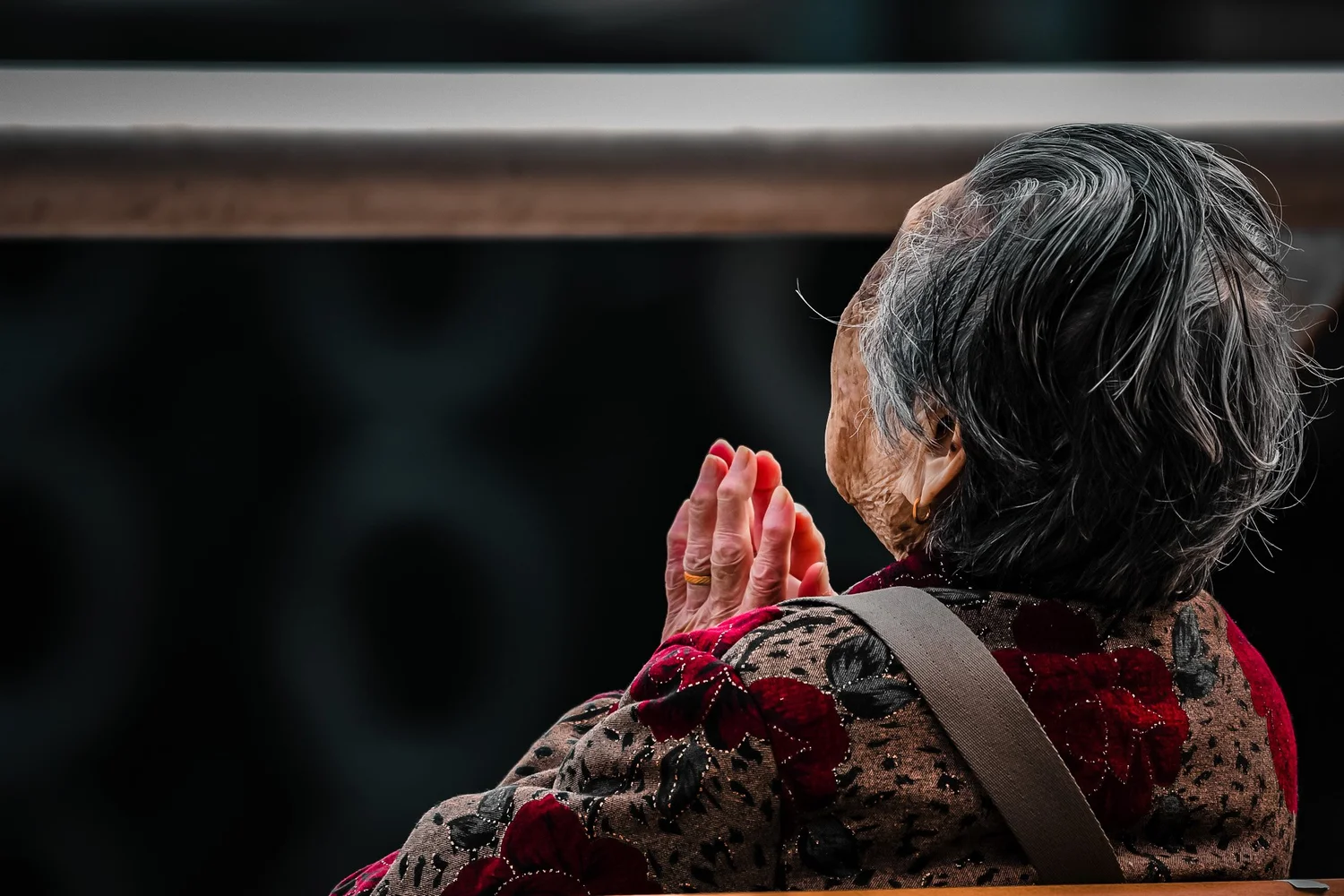 Elderly woman praying with hands pressed together in dim light, a low-light street photograph from Hong Kong