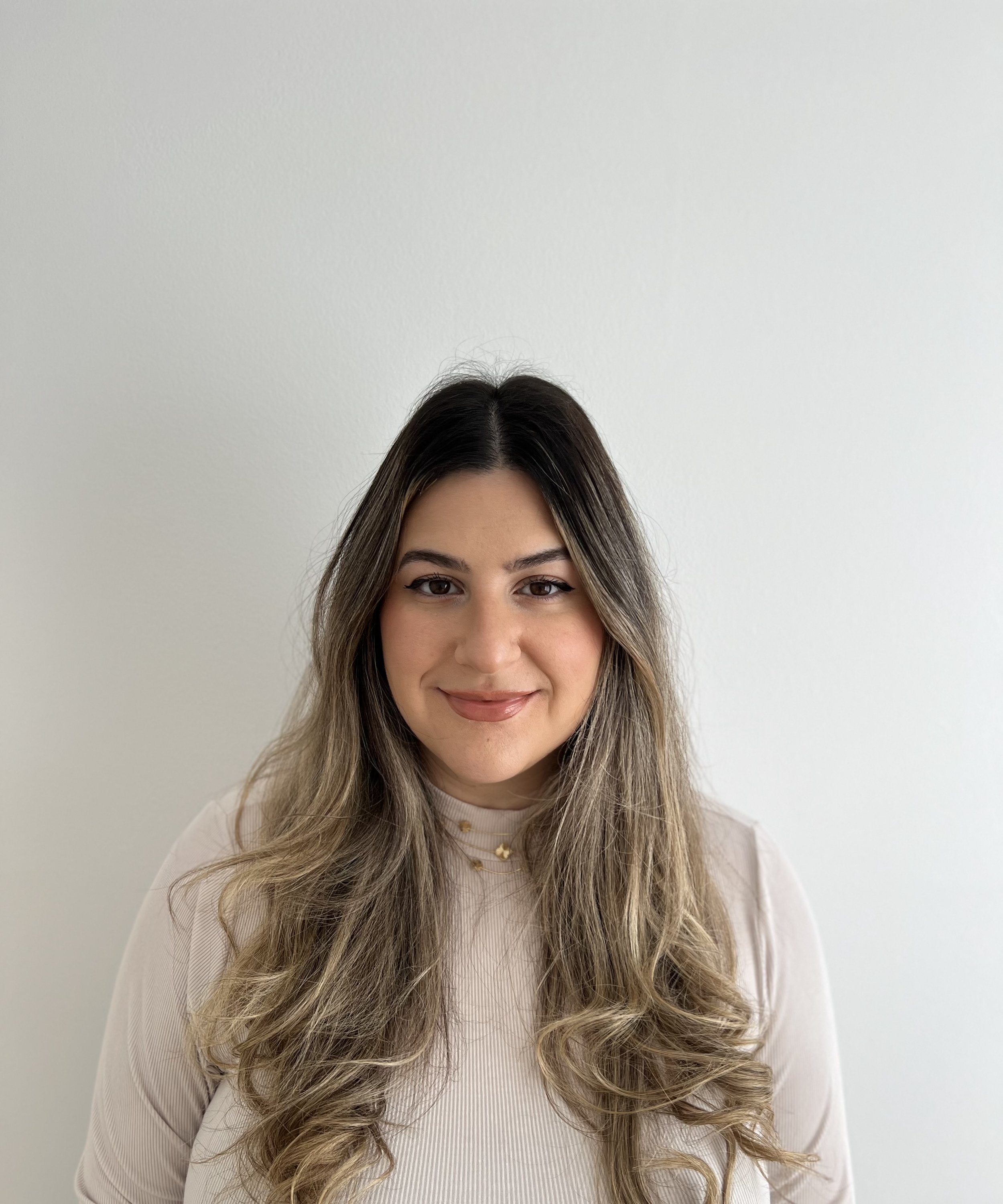 A young woman smiling with long, wavy hair, wearing a light-colored top and a small gold necklace, standing against a plain white wall.