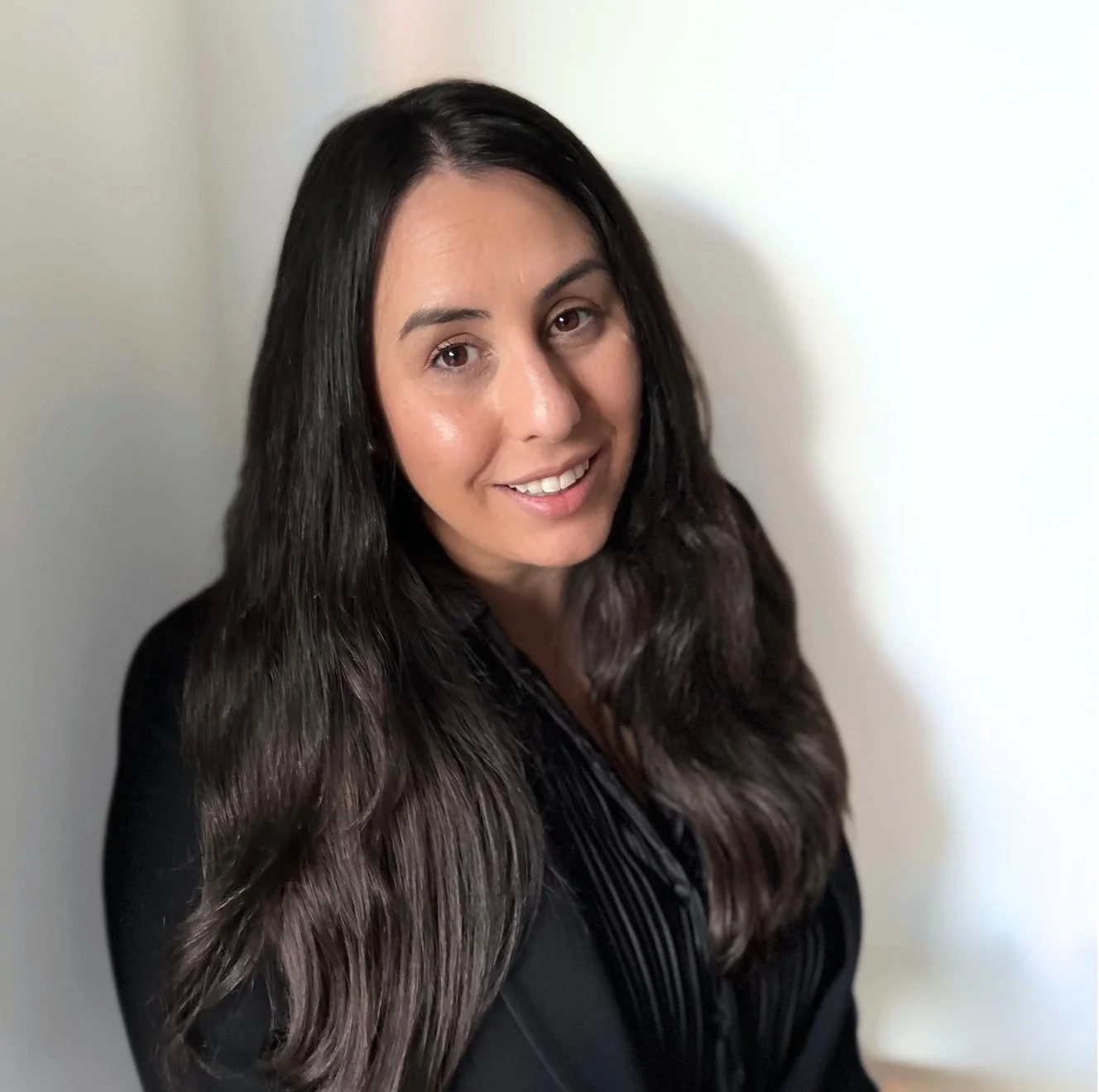 A woman with long dark hair smiling at the camera against a plain light-colored wall.
