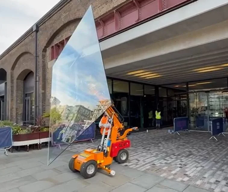 A glass panel is being installed using a small orange lift outside a building with a brick arch and glass windows.