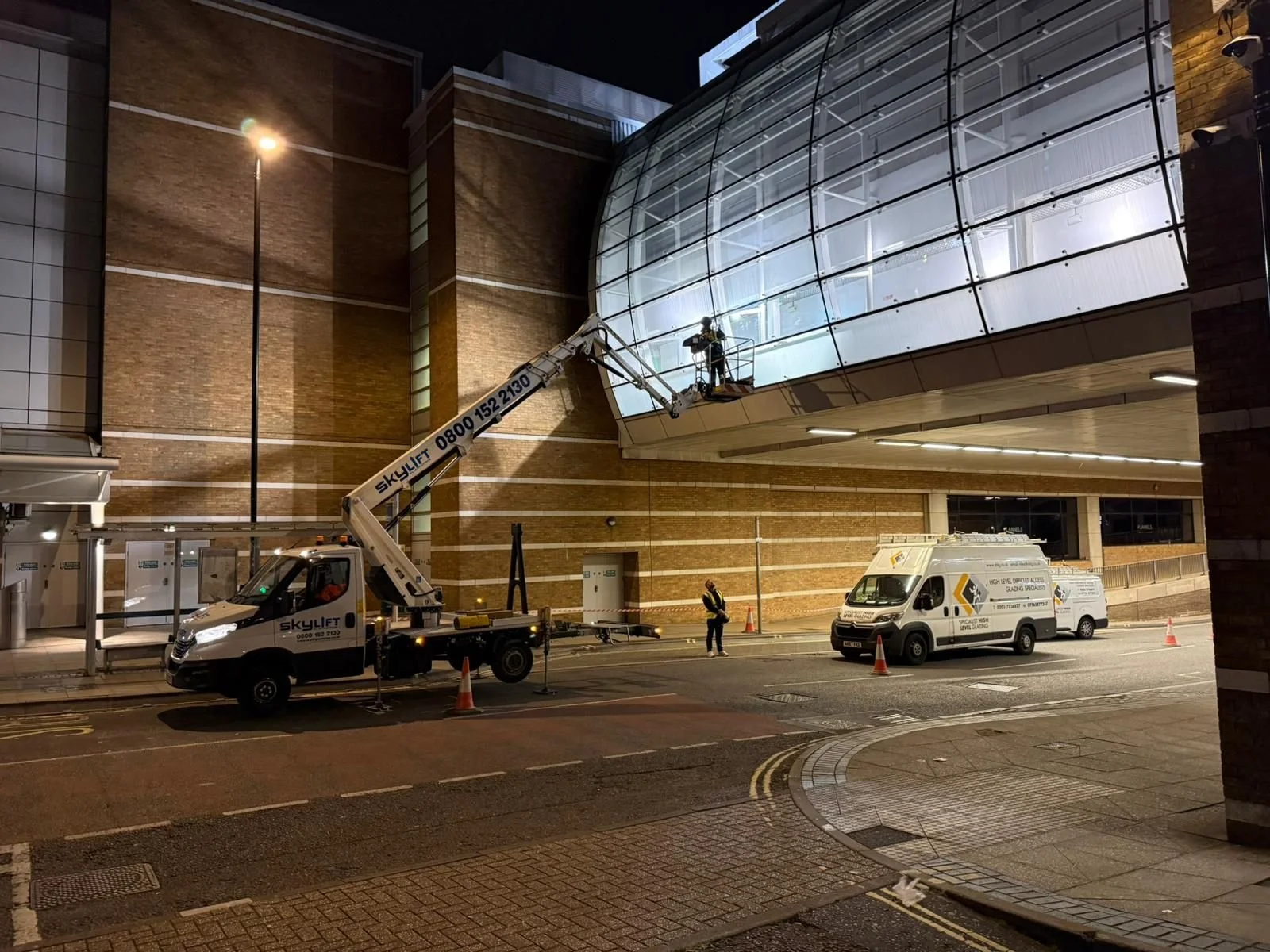 Specialise High Level Glazing Workers on a lift repairing a glass facade on a building at night, with construction cones and vehicles parked below.