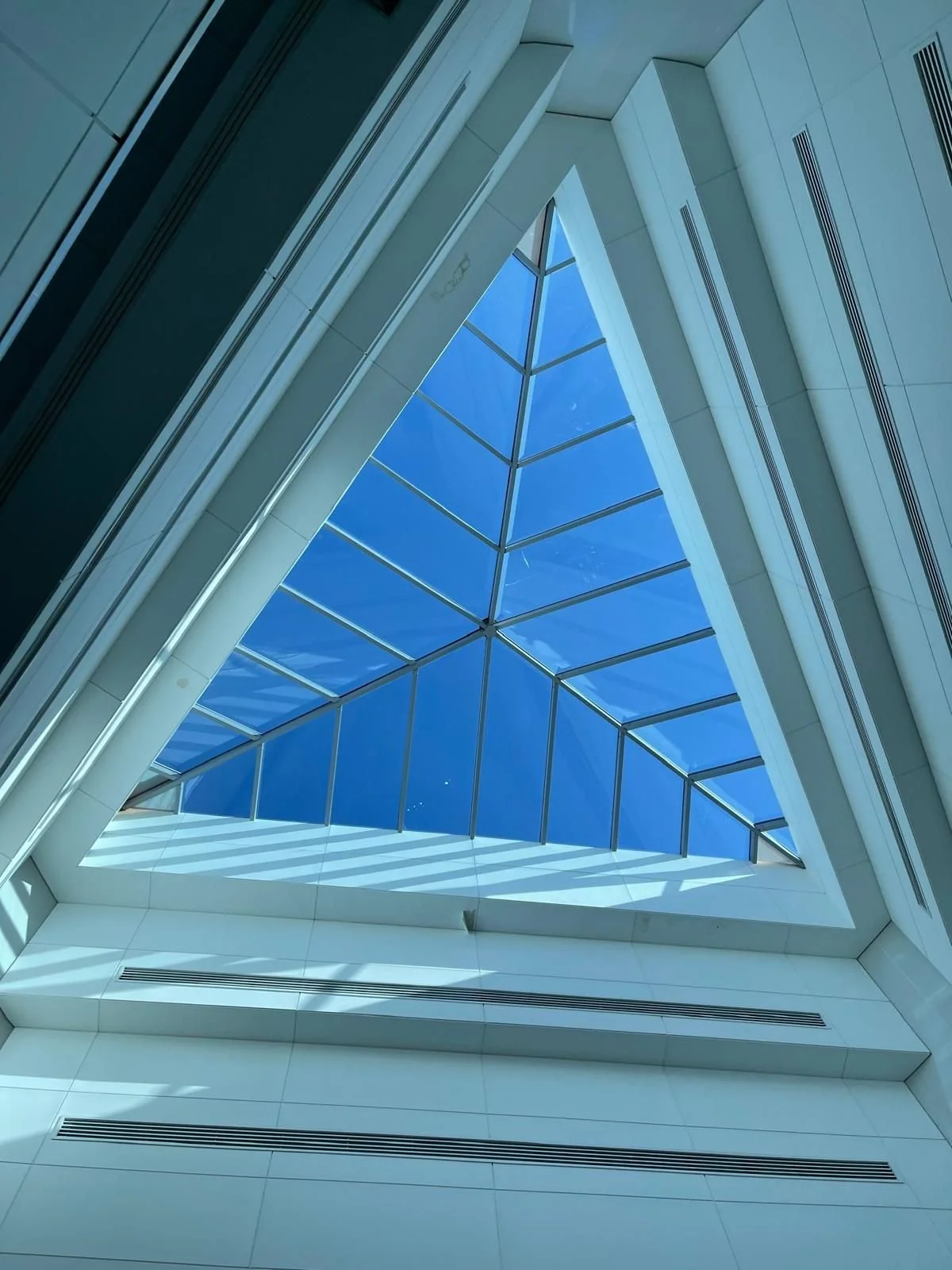 Looking up at a modern architectural ceiling with a large triangular glass skylight repair and white structural elements.