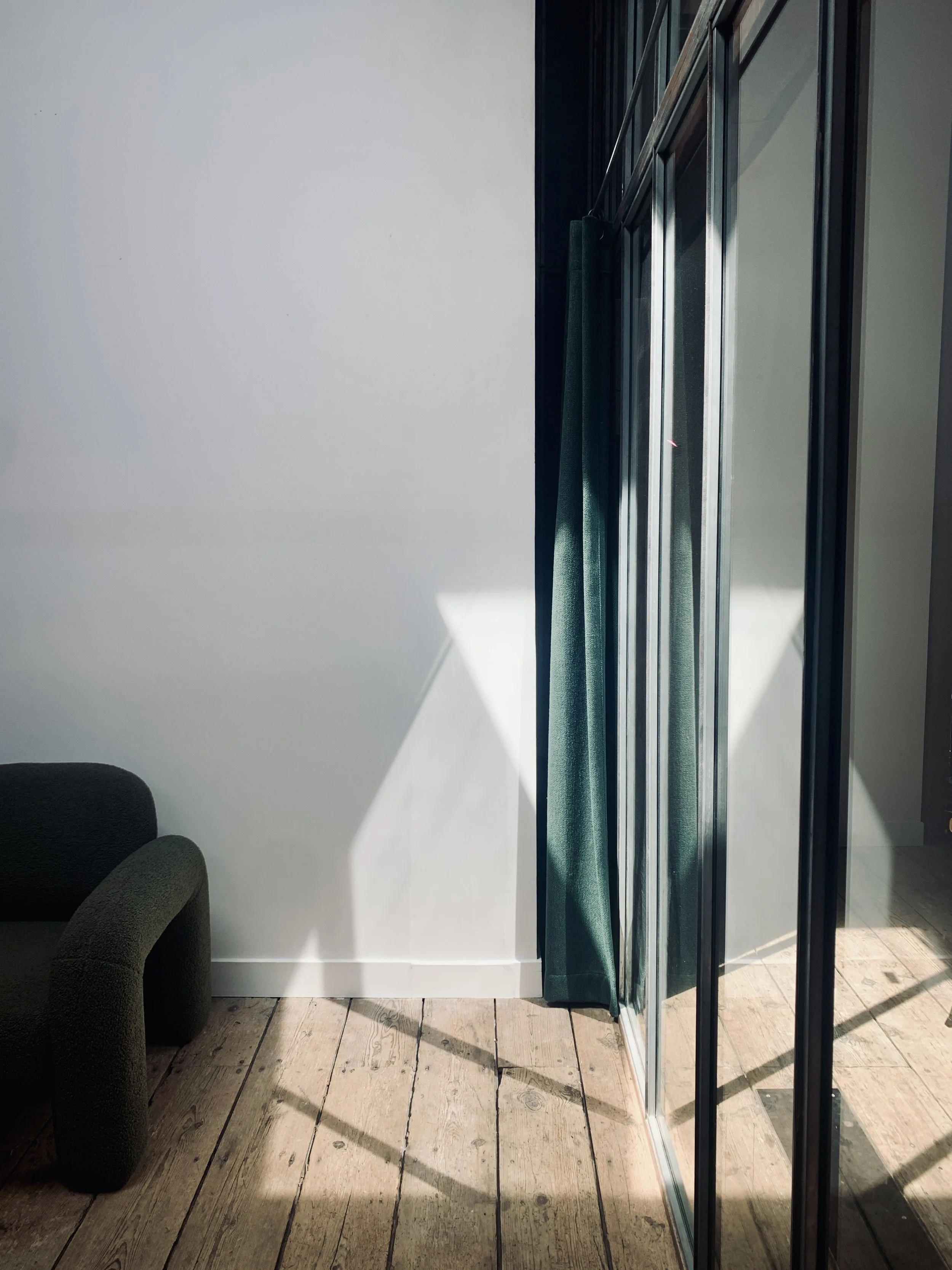 Coin salon avec un fauteuil noir, un mur blanc, grande fenêtre avec rideaux verts et sol en bois clair.