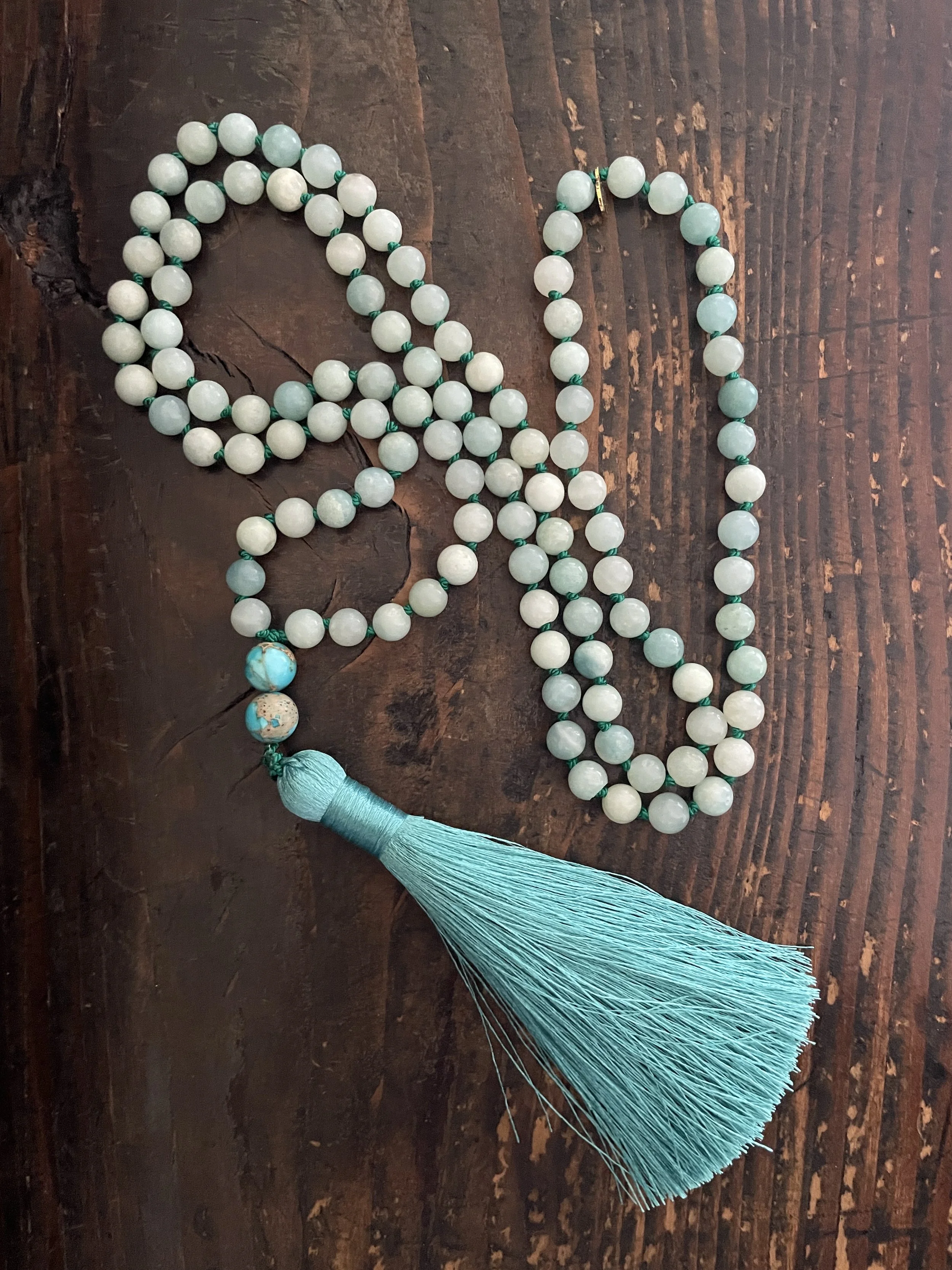 A blue amazonite beaded mala necklace with a turquoise stone and a tassel on a wooden surface.