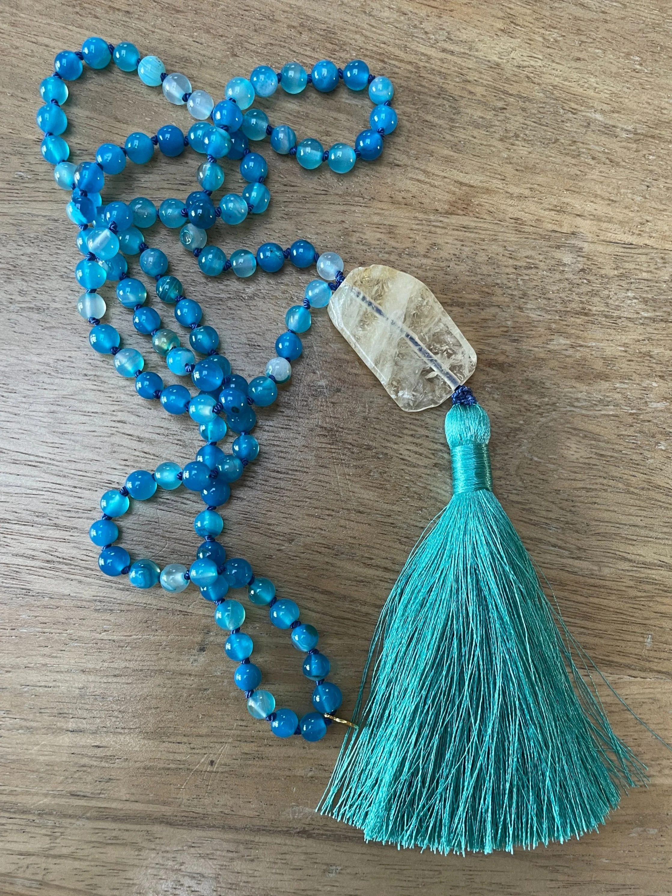 A beaded necklace with banded blue agate beads, a large citrine pendant, and a blue tassel, placed on a wooden surface.