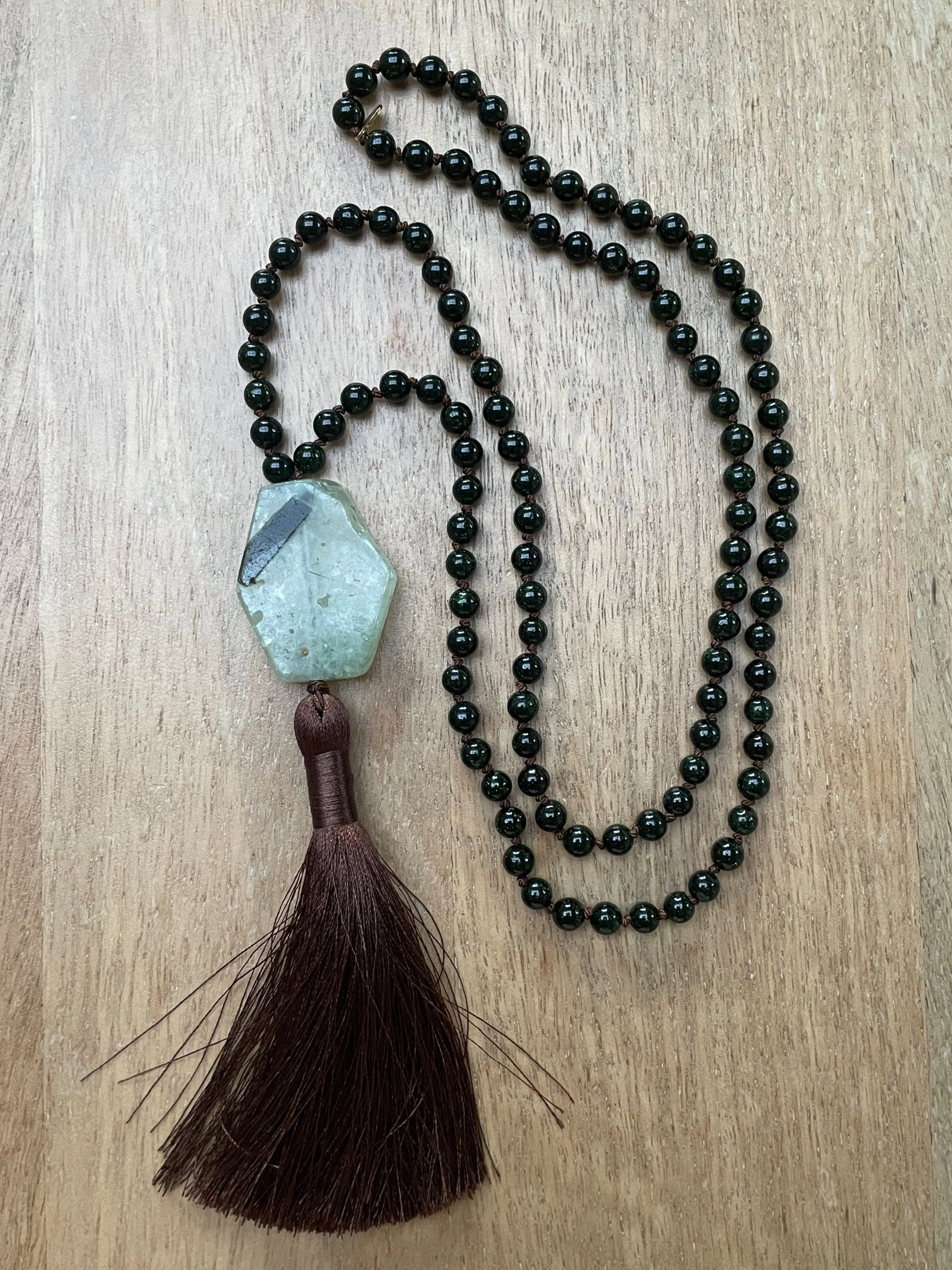 A beaded necklace with green goldstone and a large prehnite stone, a brown tassel, lying on a wooden surface.