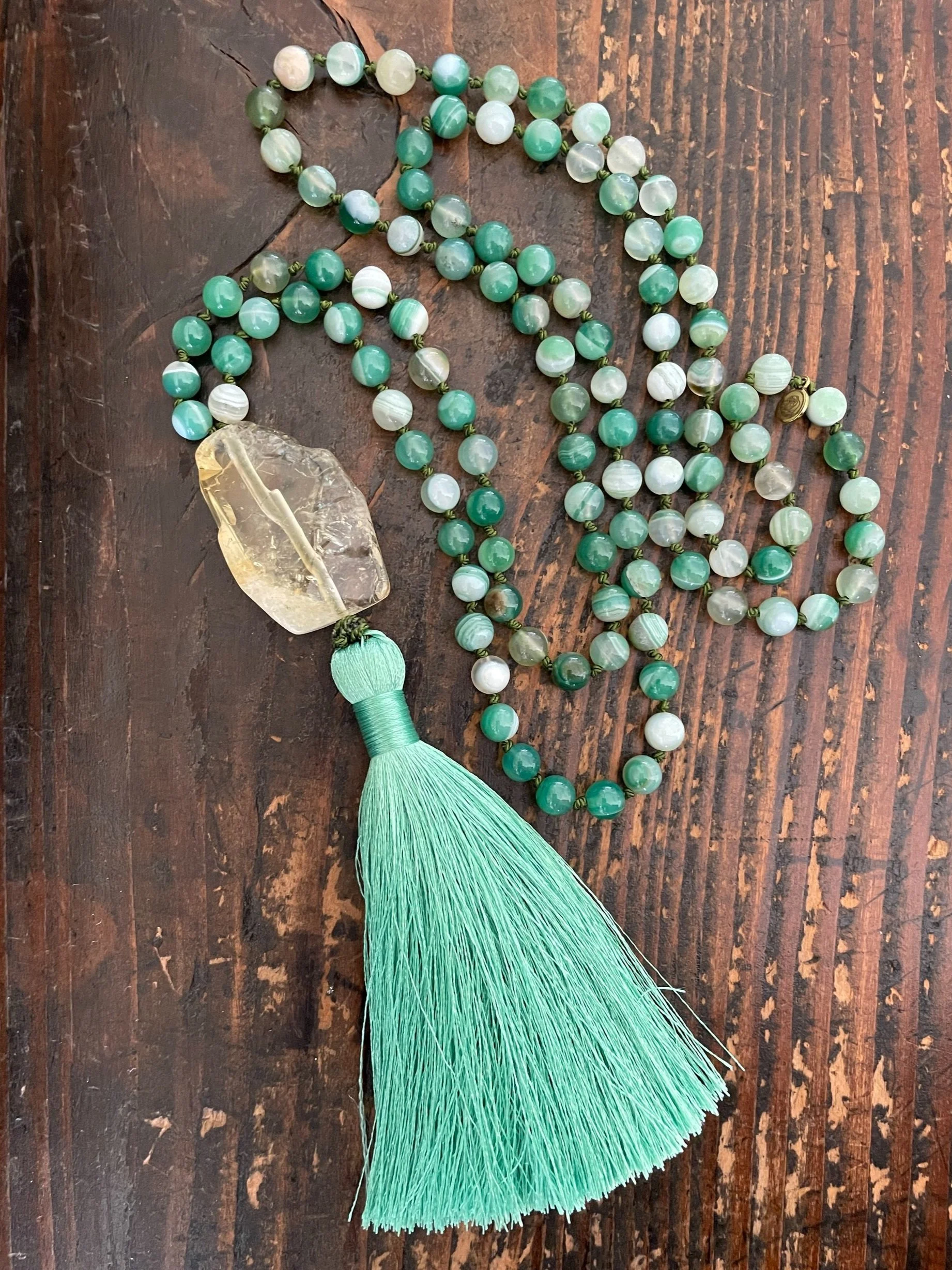 A beaded necklace with banded green agate beads, large citrine pendant, and a light green tassel, placed on a wooden surface.