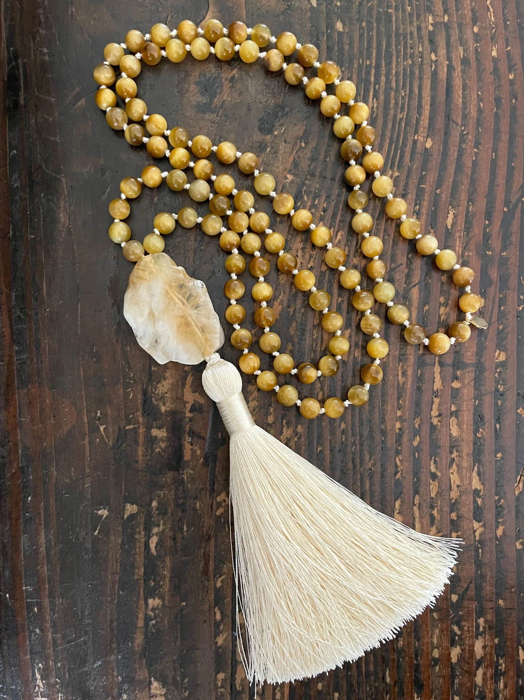A beaded tigers eye mala necklace with a large cream-colored tassel and large citrine, on a dark wooden surface.